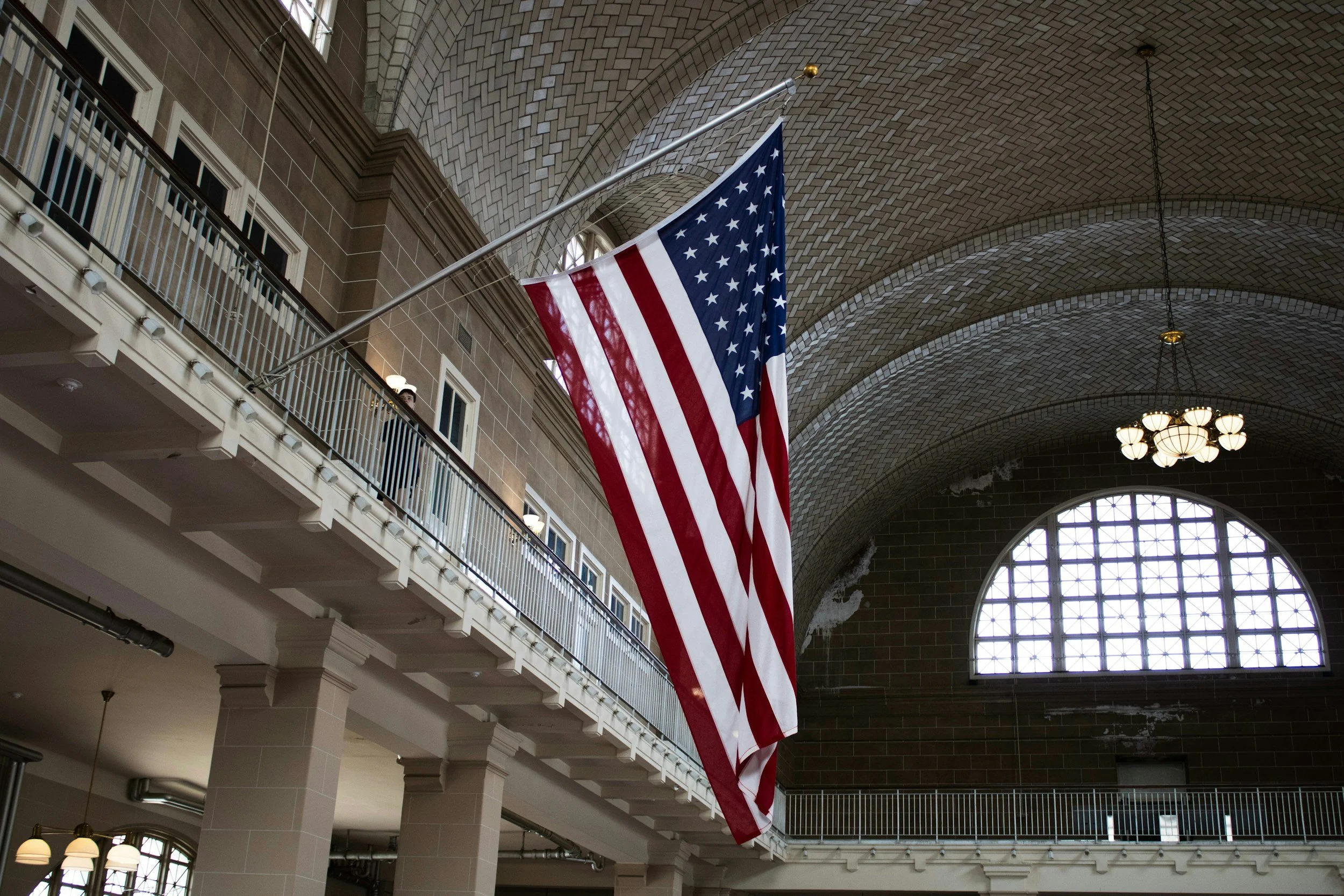 American flag hanging inside a large building with a high arched brick ceiling, chandeliers, and multiple stories with balconies and windows.