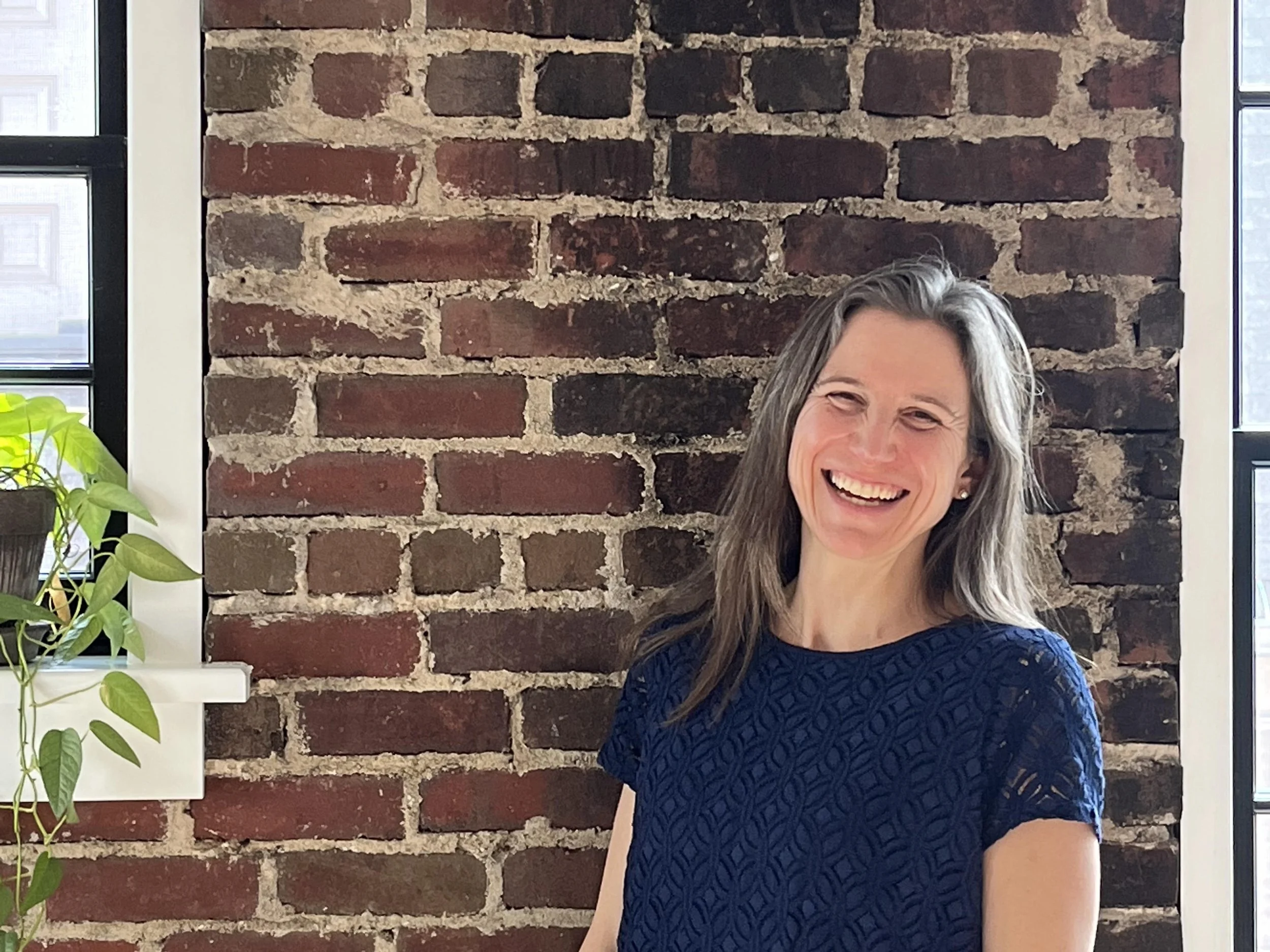 Ginny laughing in front of a brick wall with a plant on the windowsill