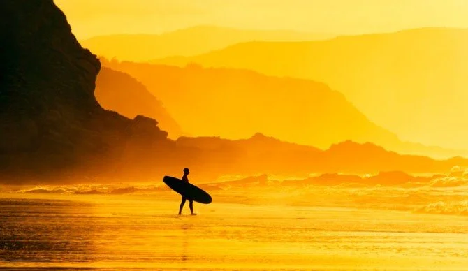 Silhouette of a person holding a surfboard walking into the ocean during sunset with a mountainous coastline in the background.