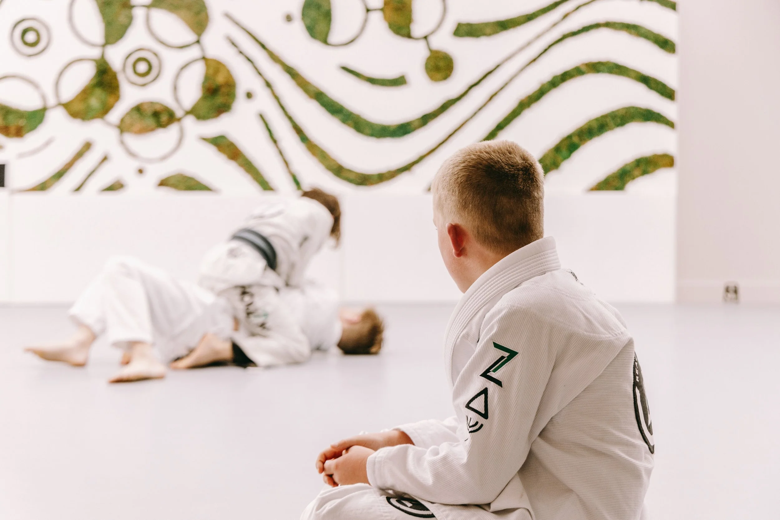 Young student in a white gi sits at the edge of the mat watching other kids practice jiu jitsu.