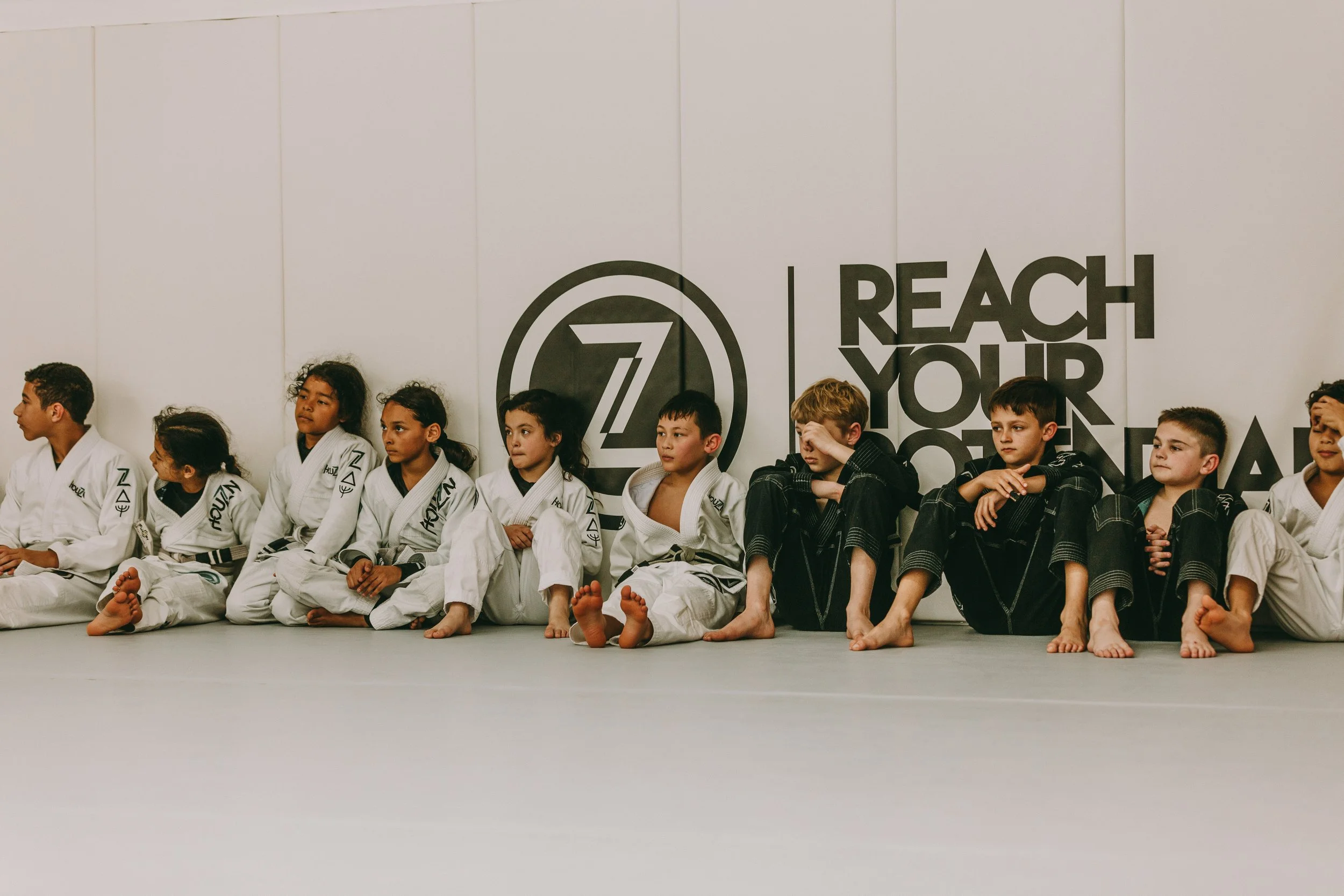 Children in white gis sit together on the mats during a kids jiu jitsu class at Houzn Jiu Jitsu Academy.