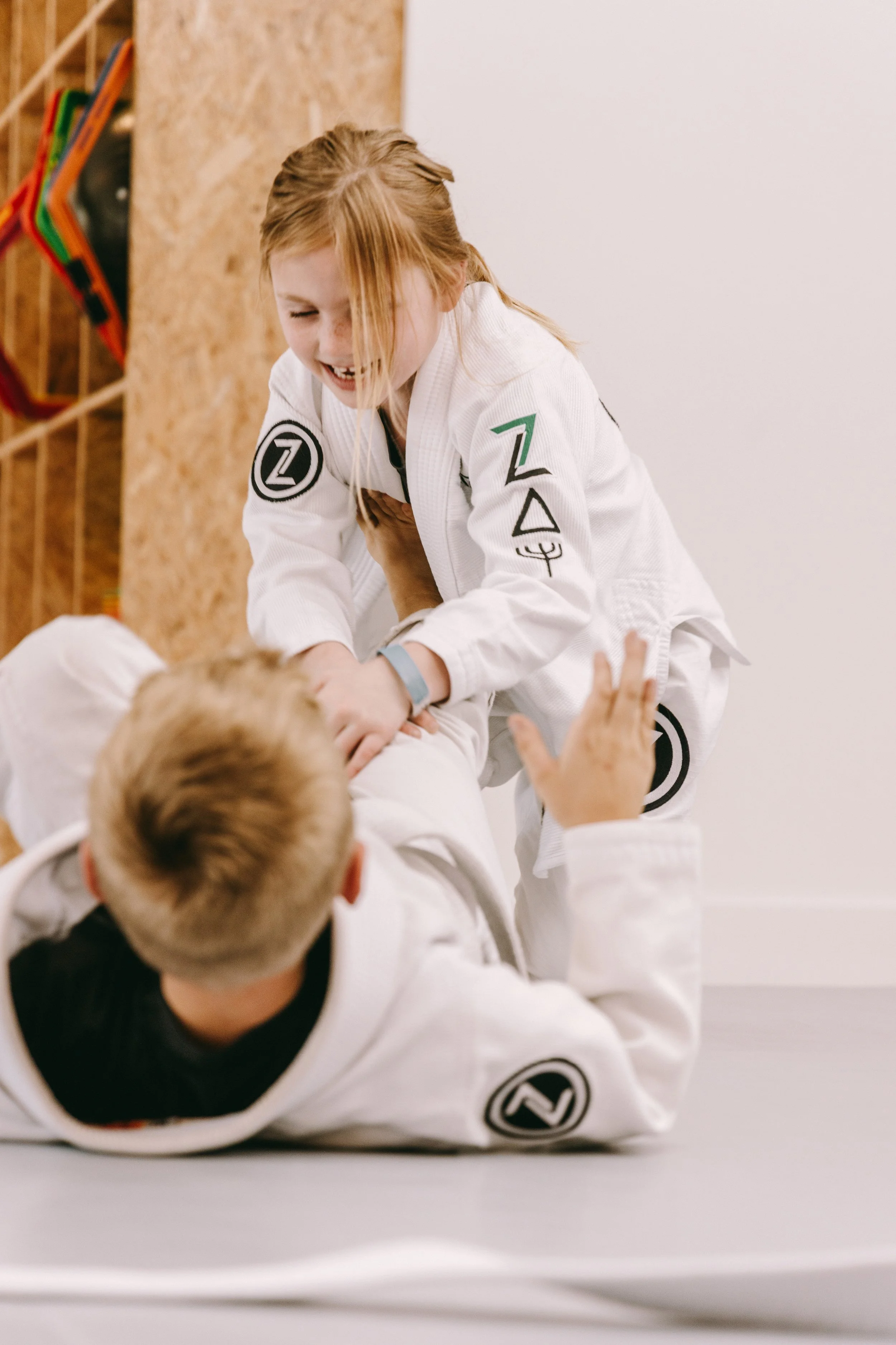 Child in a white gi smiles while practicing a Brazilian jiu jitsu drill with a partner on the mats.