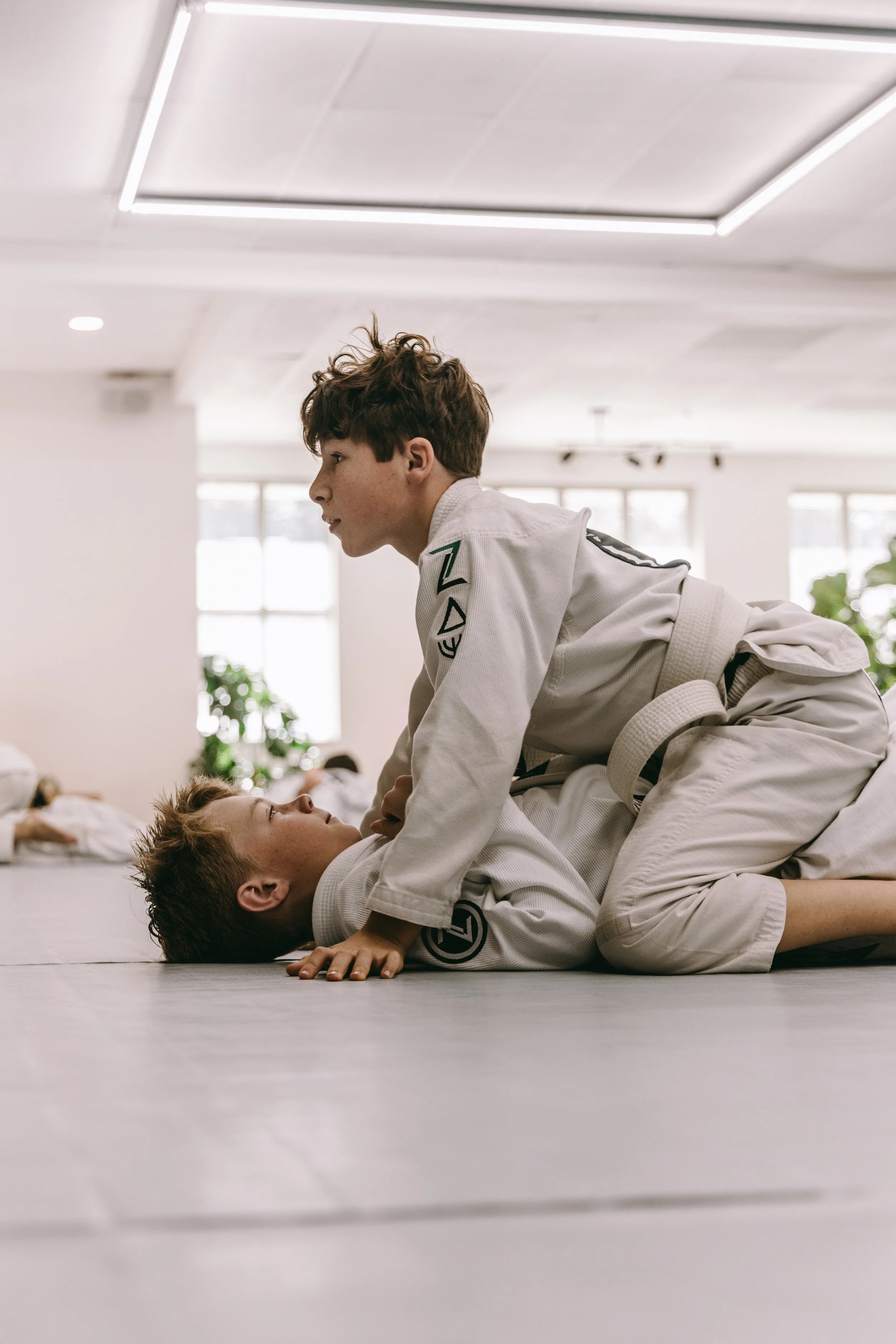 Two kids in white gis practice Brazilian jiu jitsu from the mount position during a kids class.