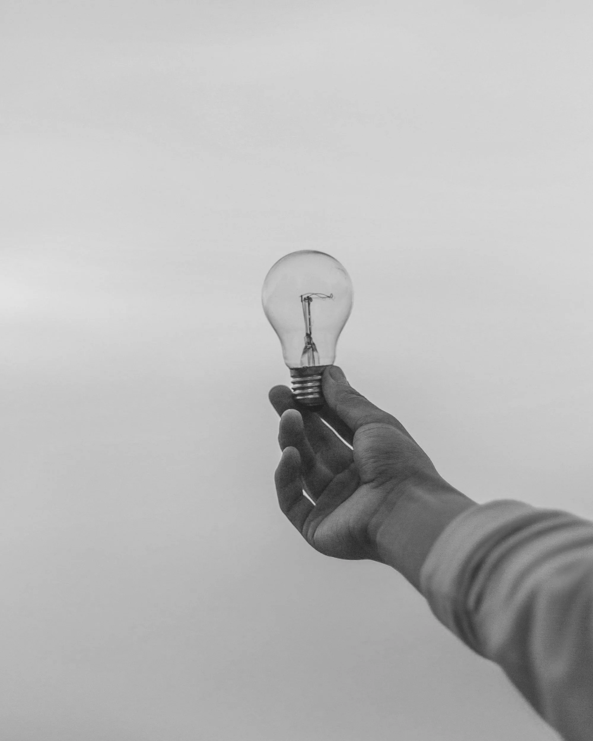 Black and white photo of a person holding a light bulb against the sky.
