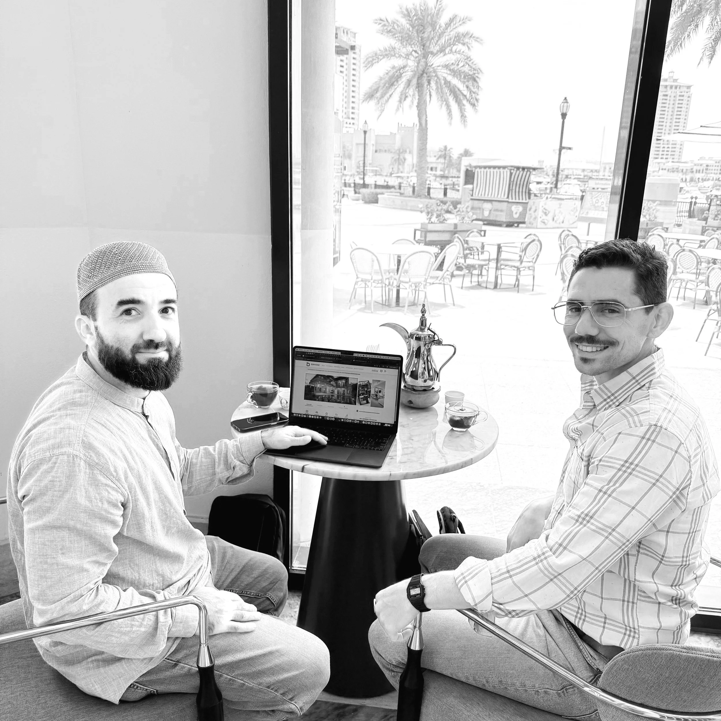 Two men sitting at a small round table inside a cafe next to a large window with outdoor seating and palm trees in the background. The man on the left has a beard, is wearing a cap and traditional clothing, and is using a laptop. The man on the right has glasses, a mustache, and is wearing a plaid shirt, smiling at the camera. There is a teapot, cups with drinks, and a paintbrush on the table.