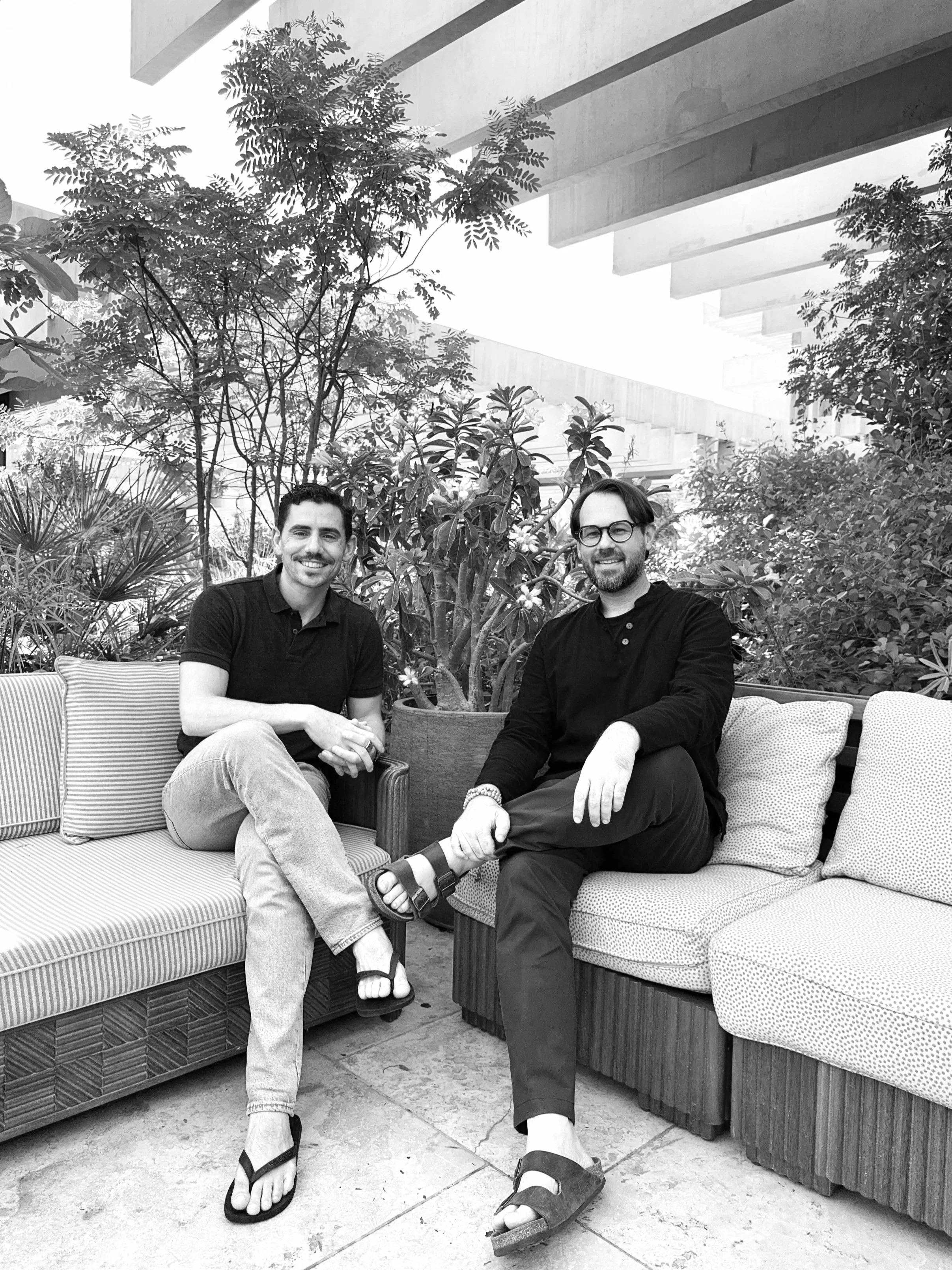Two men sitting on outdoor couches with plants and trees in the background, both smiling. One is wearing a short-sleeved shirt and jeans, and the other is in a long-sleeved shirt and pants.