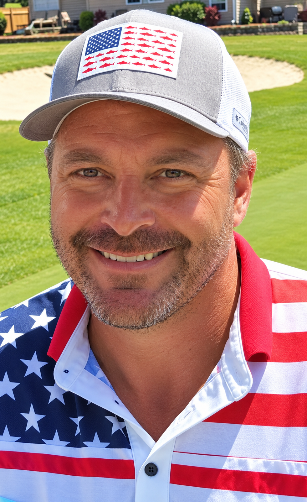 A smiling man with light on his face, wearing a gray baseball cap with an American flag patch, and a shirt featuring the American flag pattern. The background shows a grassy area and a sand trap on a golf course.