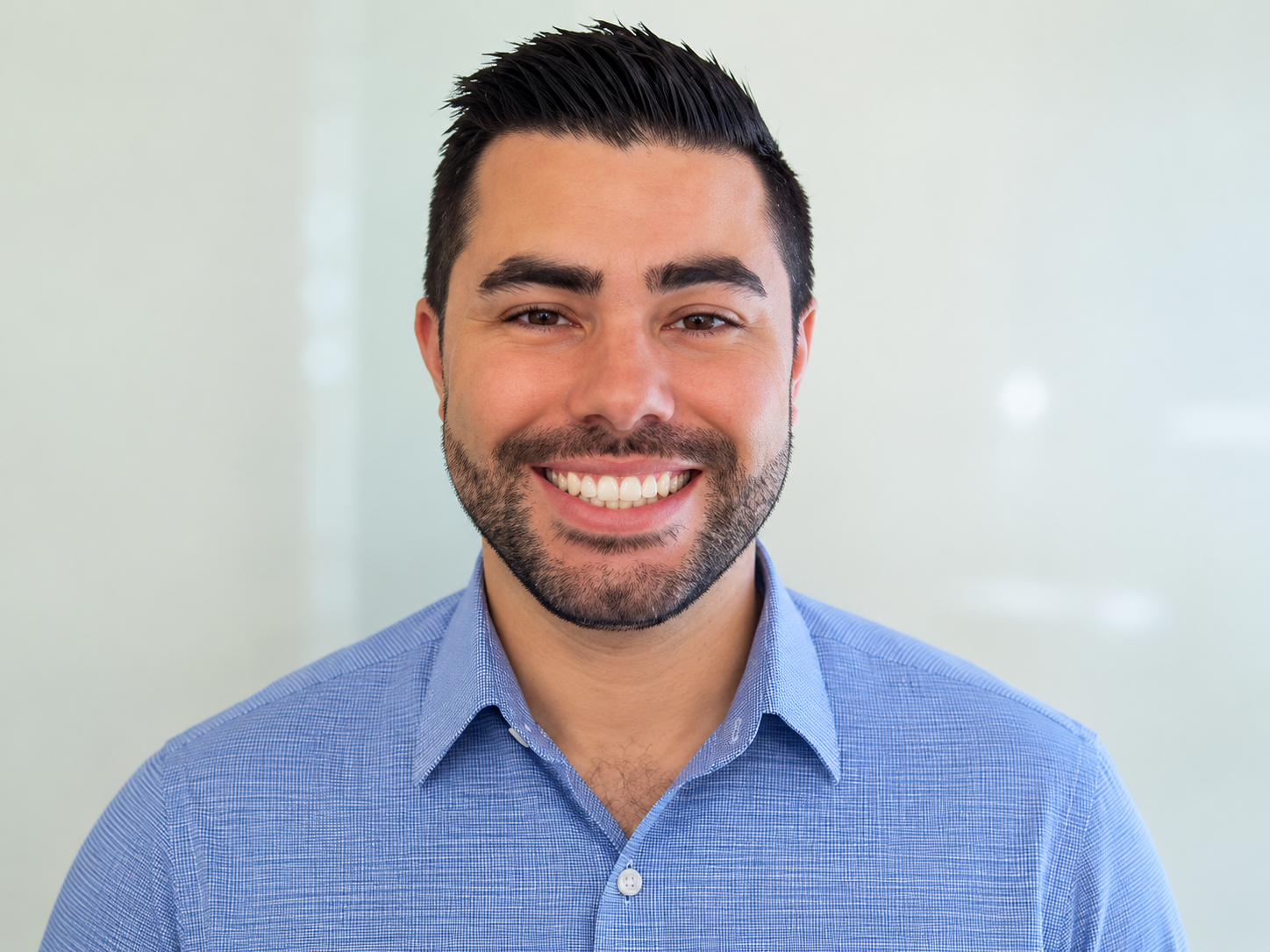 Close-up of a smiling young man with dark hair, beard, wearing a blue collared shirt, against a plain, light-colored background.