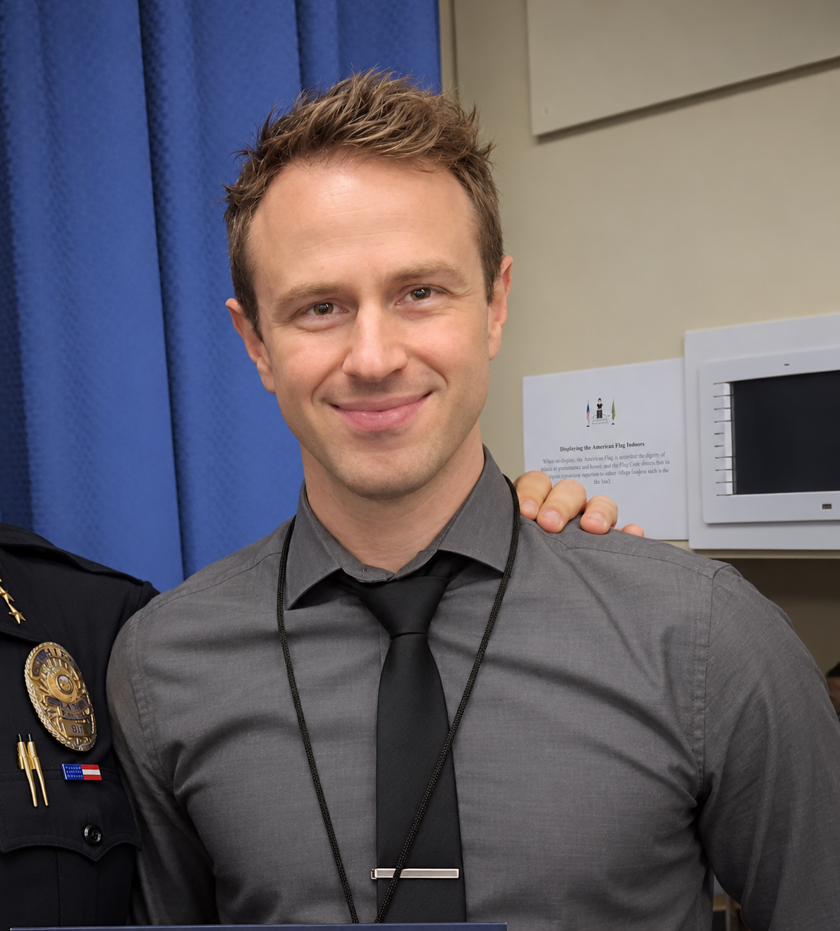 A smiling young man in a gray shirt and black tie at an indoor event, with a person in a police uniform to his left and a hand resting on his shoulder.