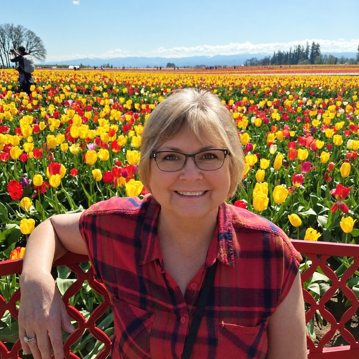 A woman with glasses smiling, wearing a red plaid shirt, sitting in front of a colorful tulip field on a sunny day.