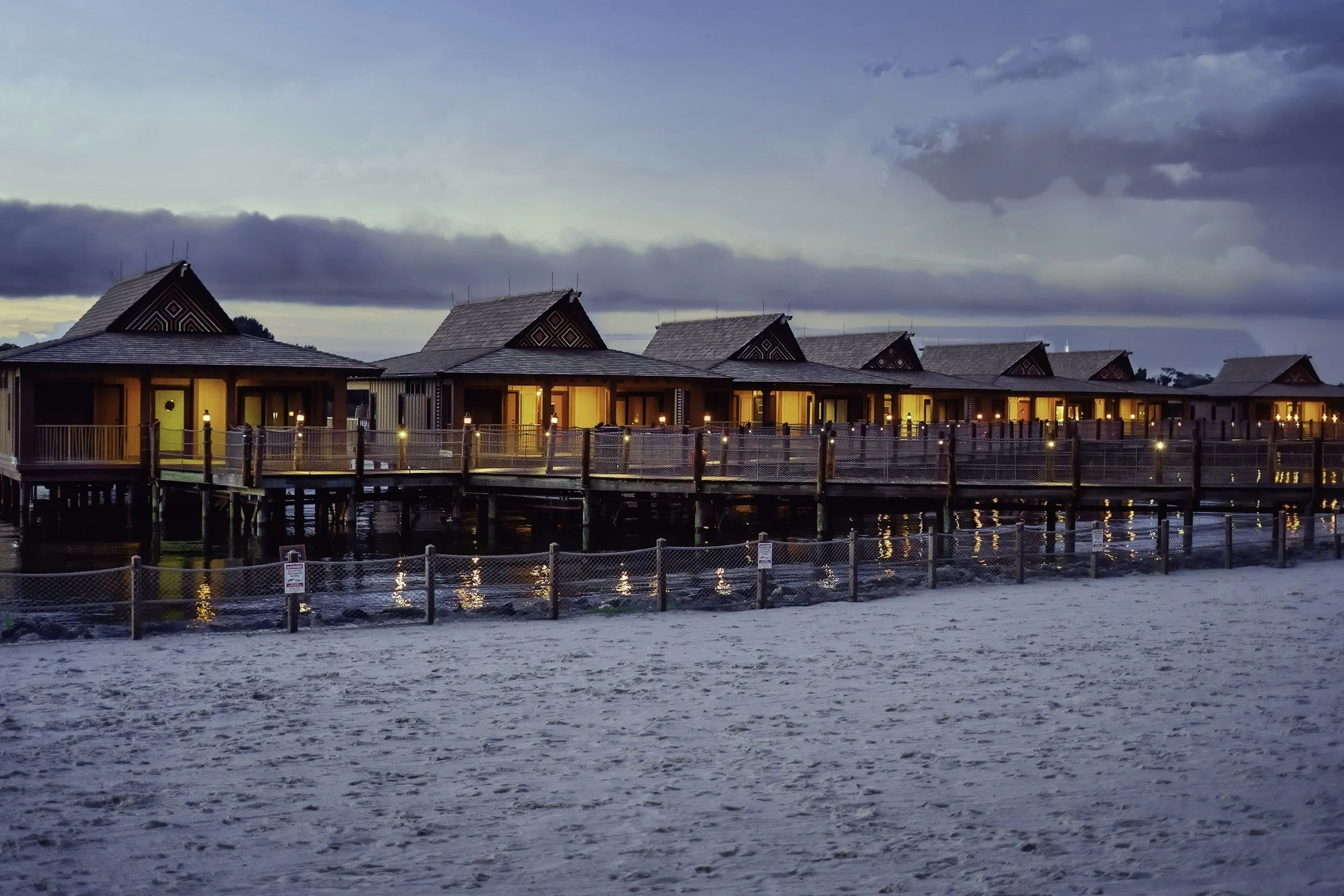 Overwater bungalows with lit windows at dusk, overlooking a sandy beach with fencing and cloudy sky