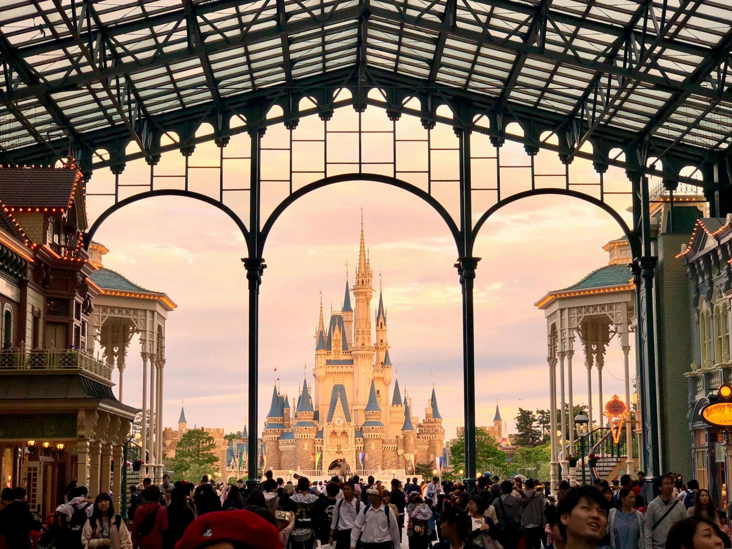 Cinderella Castle at Disney theme park viewed from inside a covered walkway, with many visitors walking and taking photos at sunset.