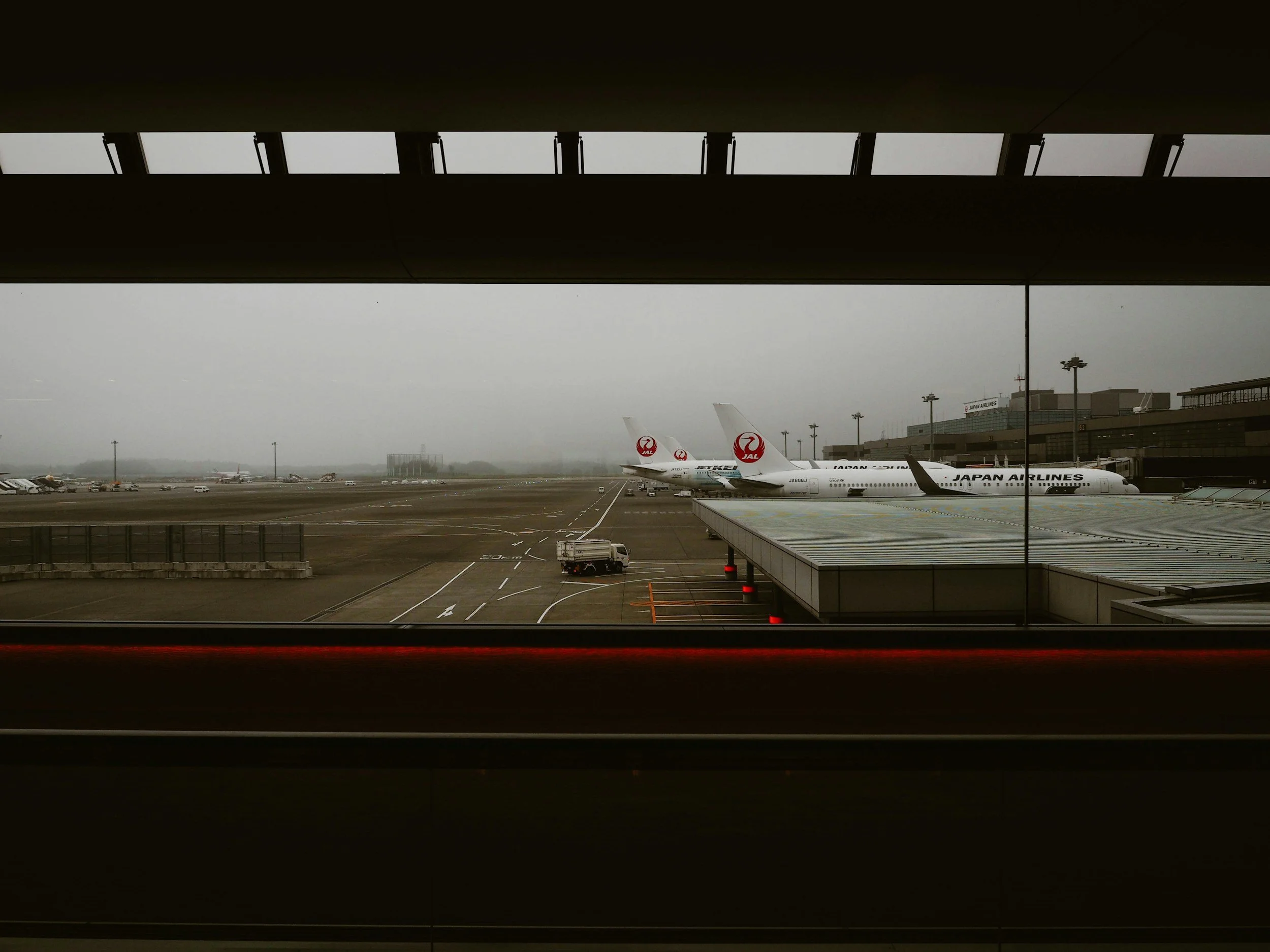 View of an airport tarmac with Japan Airlines airplanes parked at the terminal, seen through large window with structural beams.