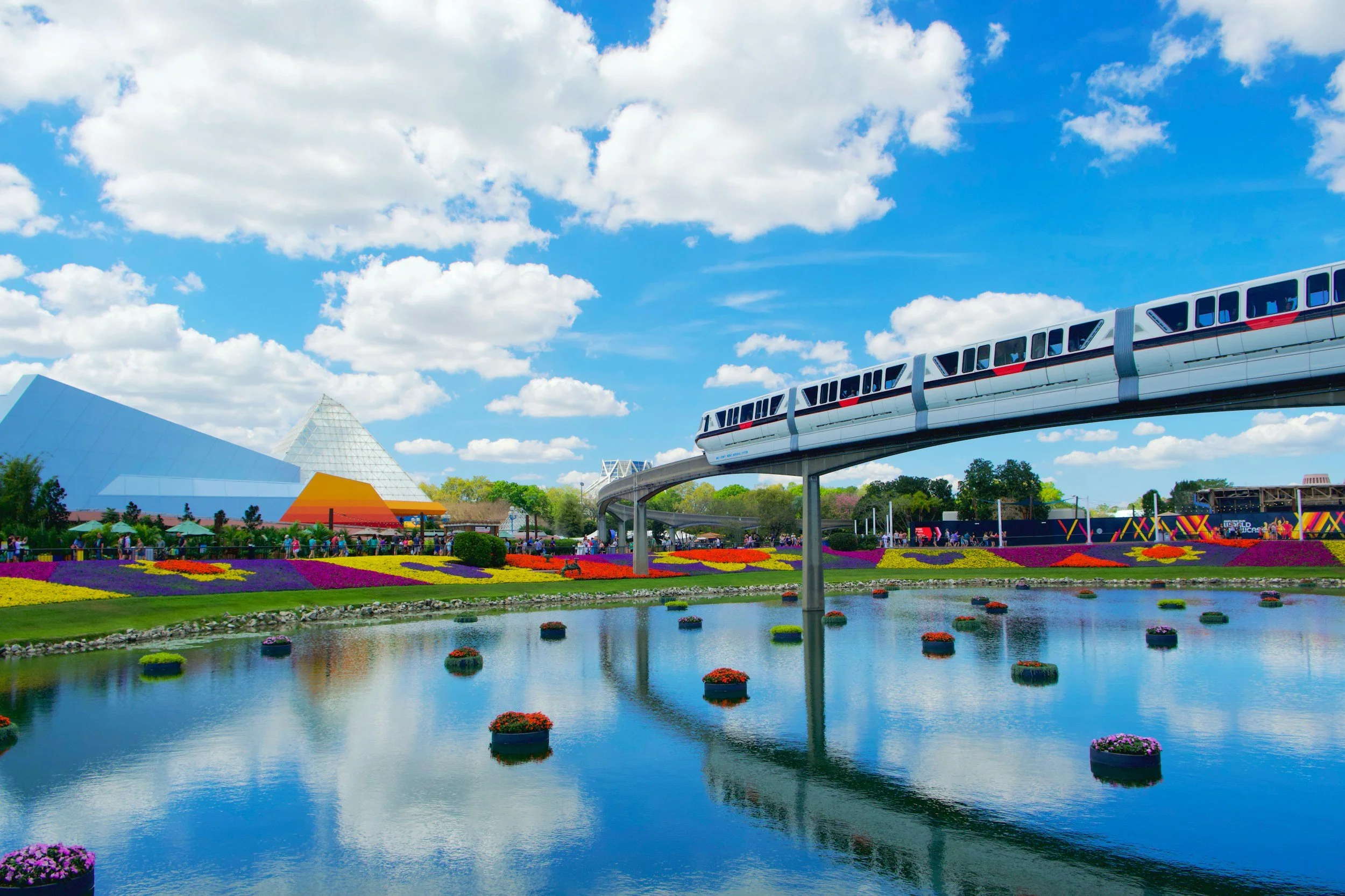 Epcot theme park at Walt Disney World, showing a monorail train traveling above a pond with floating flower baskets, colorful flowerbeds, and the iconic Geodesic Sphere of Spaceship Earth in the background under a partly cloudy blue sky.