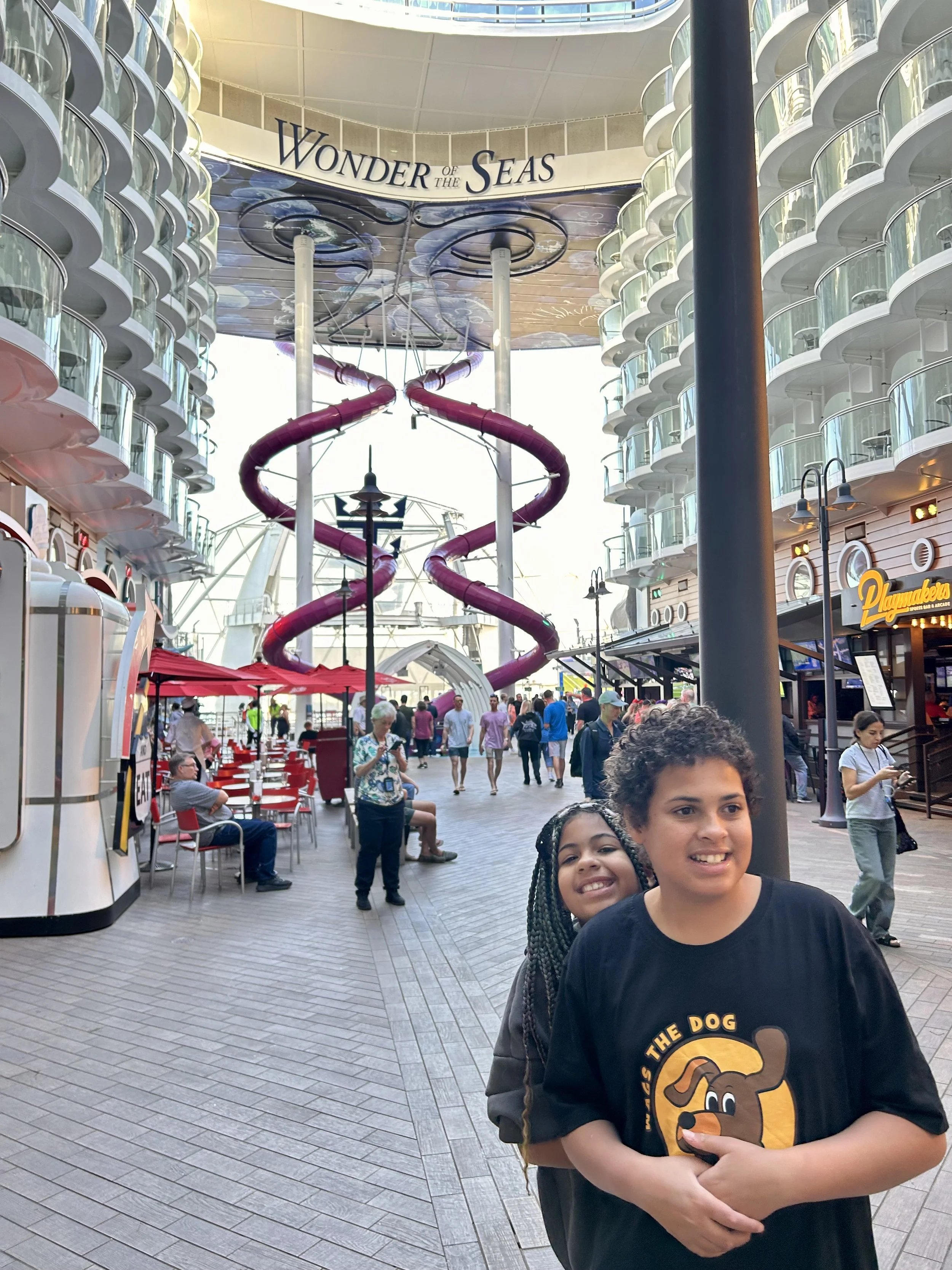 Two children standing on a cruise ship promenade, smiling, with the Wonder of the Seas cruise ship's water slide and balconies in the background.