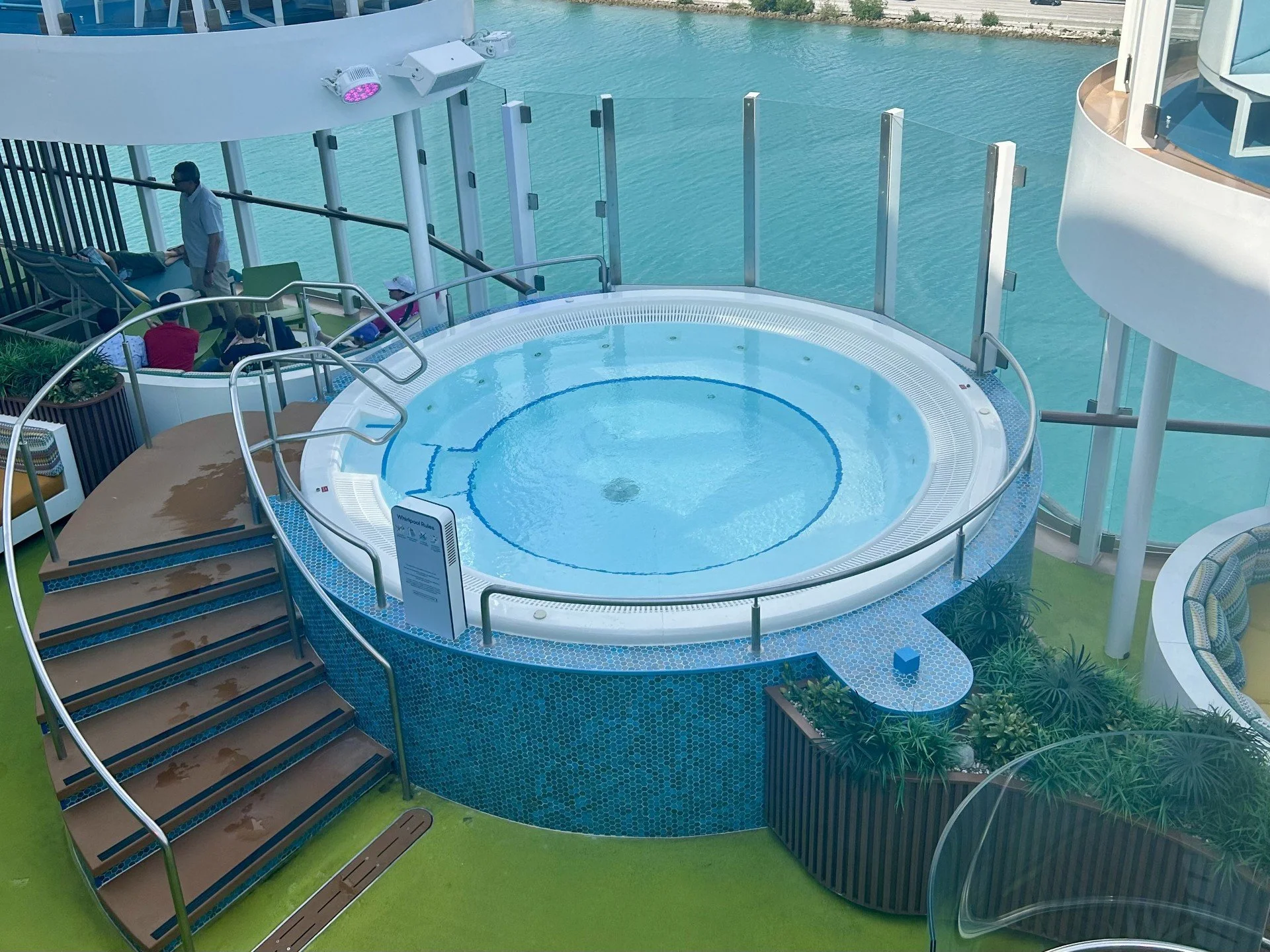 A small round hot tub with blue tiles around the exterior, located on a deck overlooking water. There are stairs leading up to the hot tub, with a railing surrounding it. Nearby, there are lounge chairs, and a few people are seated or standing on the deck.