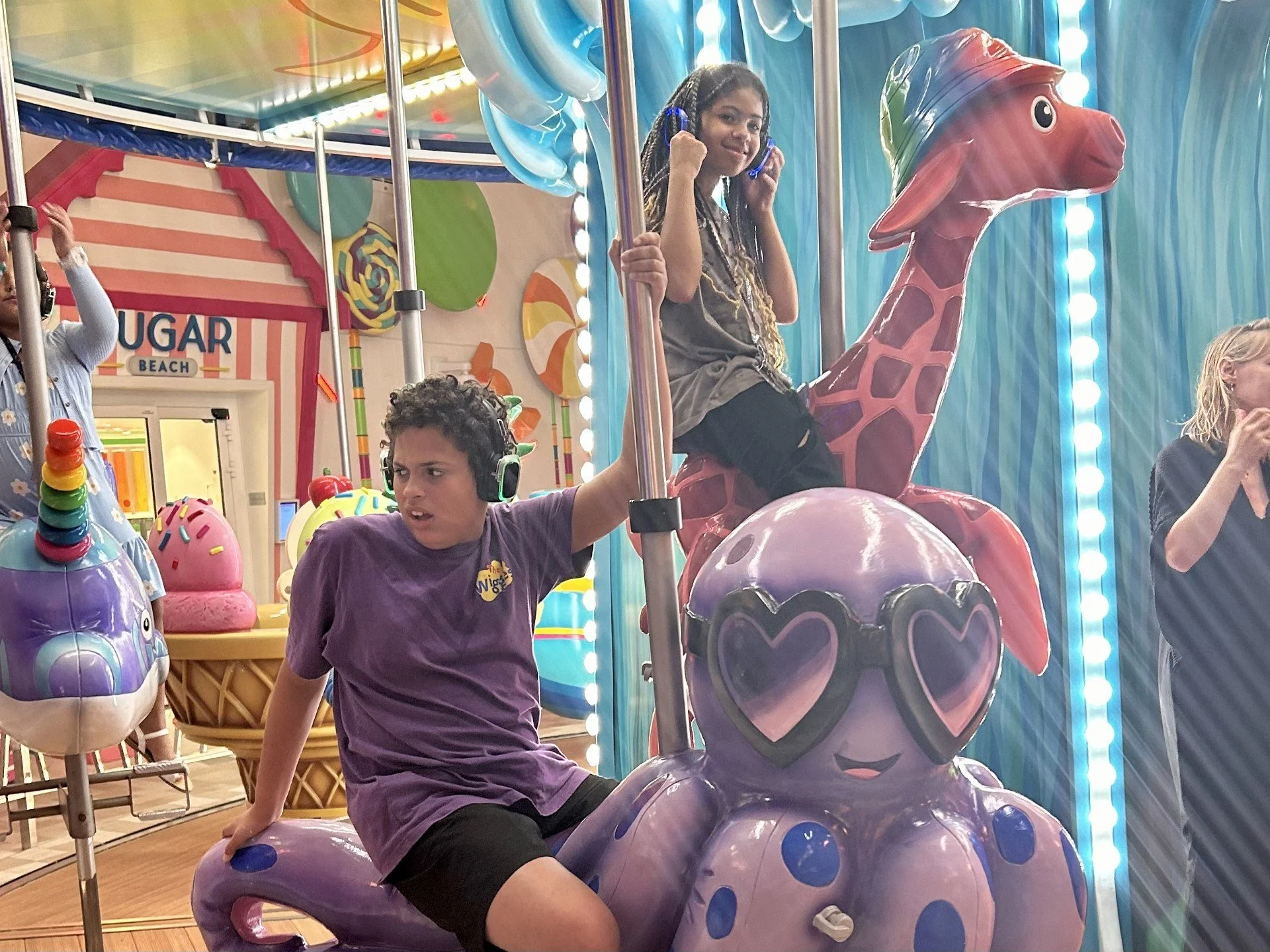 Children enjoying a colorful amusement ride with a purple octopus wearing heart-shaped sunglasses and a pink giraffe for a part of the ride, in an arcade or amusement park.
