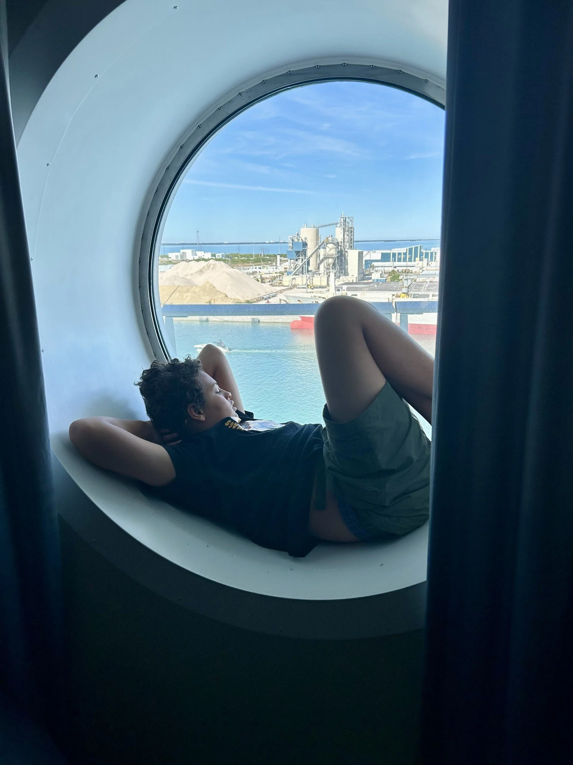 A young boy lies inside a space capsule window, looking out at an industrial port with water, ships, and industrial buildings under a blue sky.