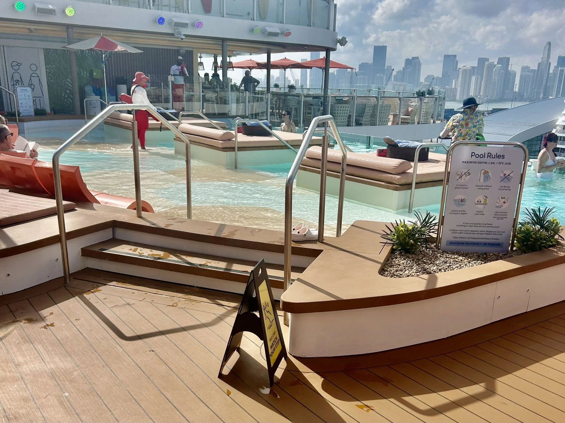 Rooftop pool area with pool rules sign, lounge chairs, and city skyline in the background. People are enjoying the pool, some sitting on loungers, and a staff member standing near the pool.