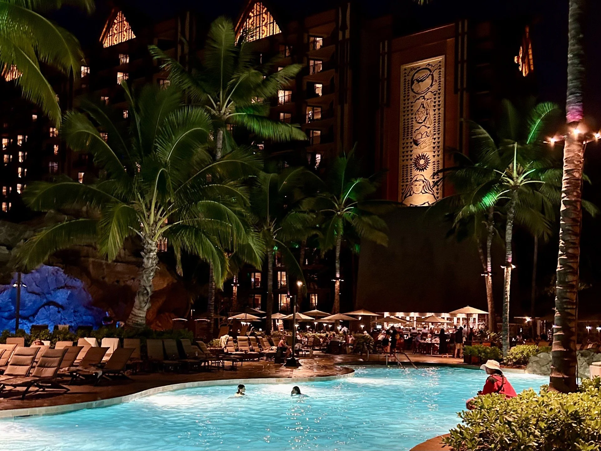 Night view of a hotel pool area with people swimming, surrounded by lounge chairs and umbrellas. Tall palm trees and an illuminated building with balconies and a decorative mural are in the background. Night time at Aulani