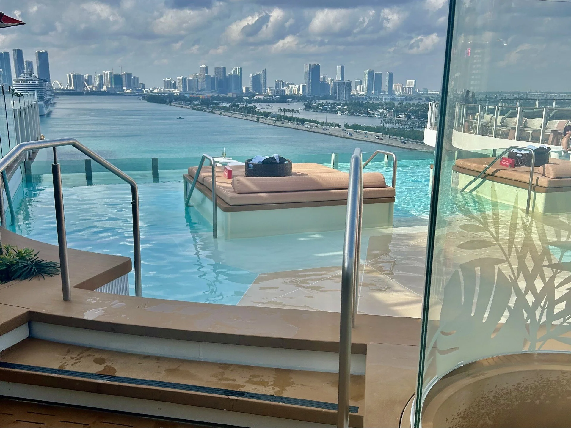 Luxury rooftop infinity pool with lounge chairs and a view of a city skyline, water, and river in the background.