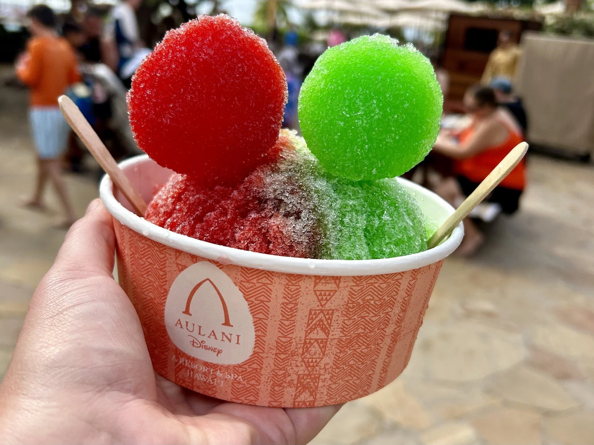 Hand holding a cup of colorful snow cones with three scoops of red and green shaved ice, labeled 'Aulani Disney Resort & Spa, Hawaii,' with blurred people in background.