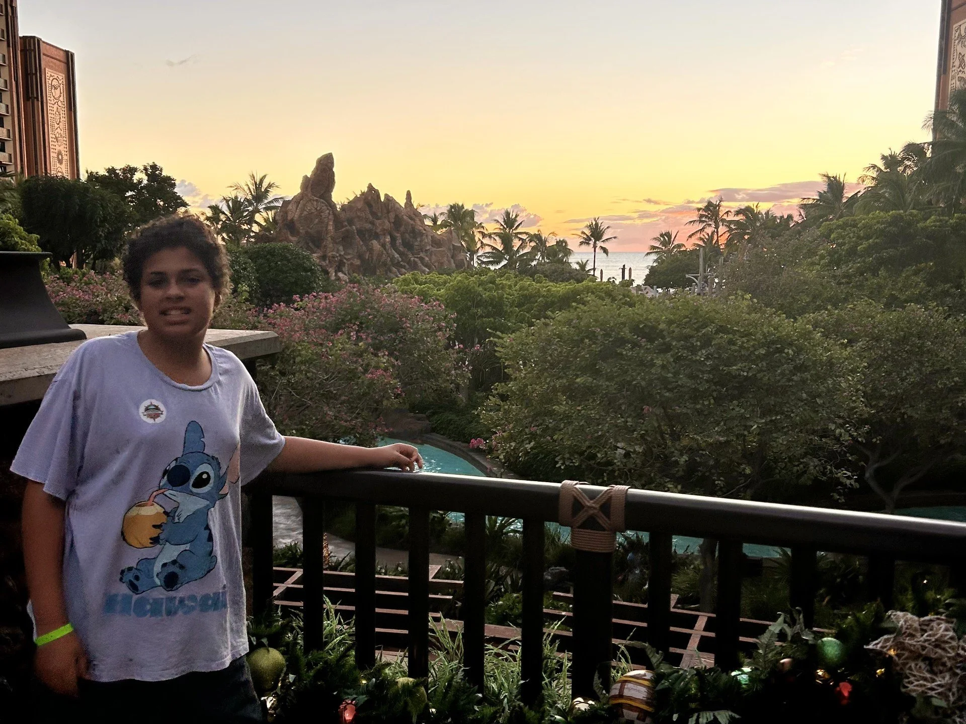 A young boy with curly hair wearing a Stitch from Lilo & Stitch t-shirt and a yellow wristband standing outside on a balcony during sunset. The background features lush tropical vegetation, pink flowers, at Aulani in Oahu