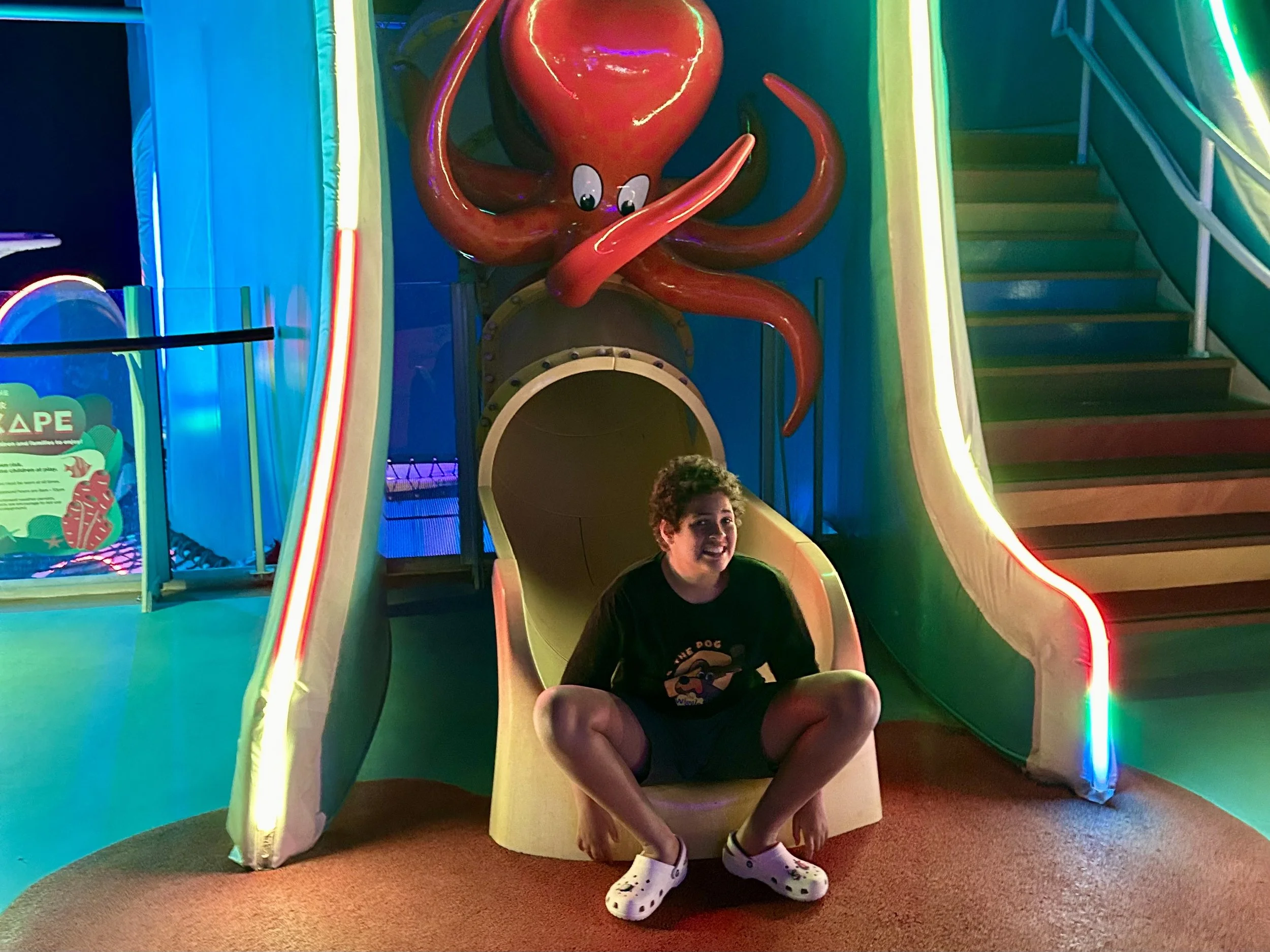 A boy sitting at the bottom of a small yellow slide, which is part of an indoor play structure shaped like a squid with pink tentacles and cartoon eyes, in a brightly lit indoor play area with colorful neon lighting.