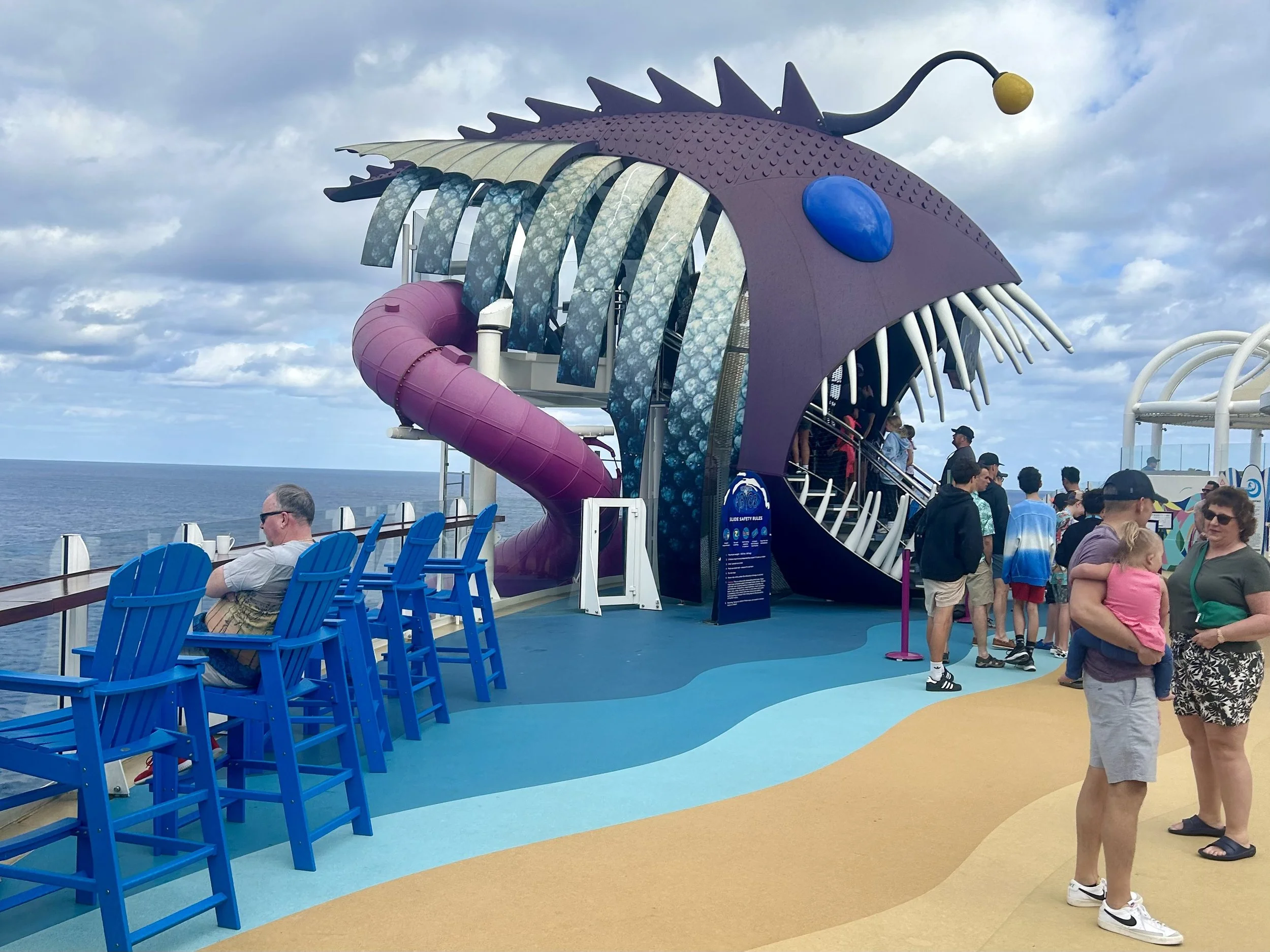 People on a cruise ship deck with a large colorful fish-themed waterslide featuring a purple head, blue eyes, white teeth, and a pink tail, with the ocean and sky in the background. Ultimate Abyss Wonder of the Seas