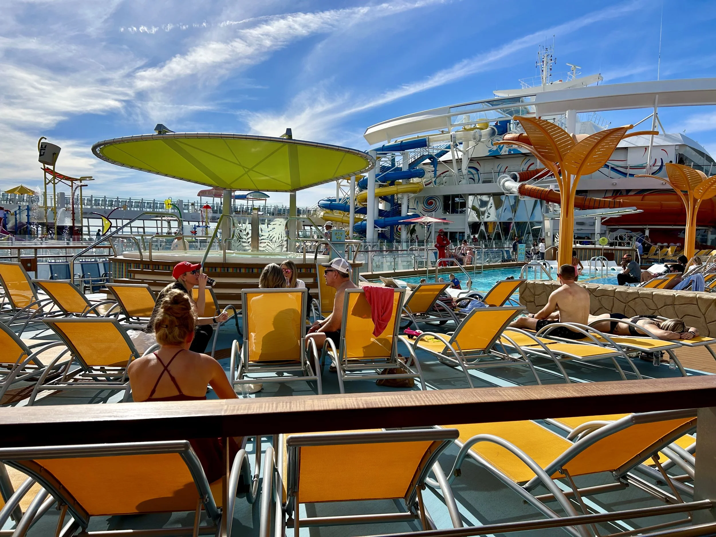 Passengers lounging on yellow deck chairs around a swimming pool, with waterslides and water attractions on a cruise ship in the background under a partly cloudy blue sky. Wonder of the Seas