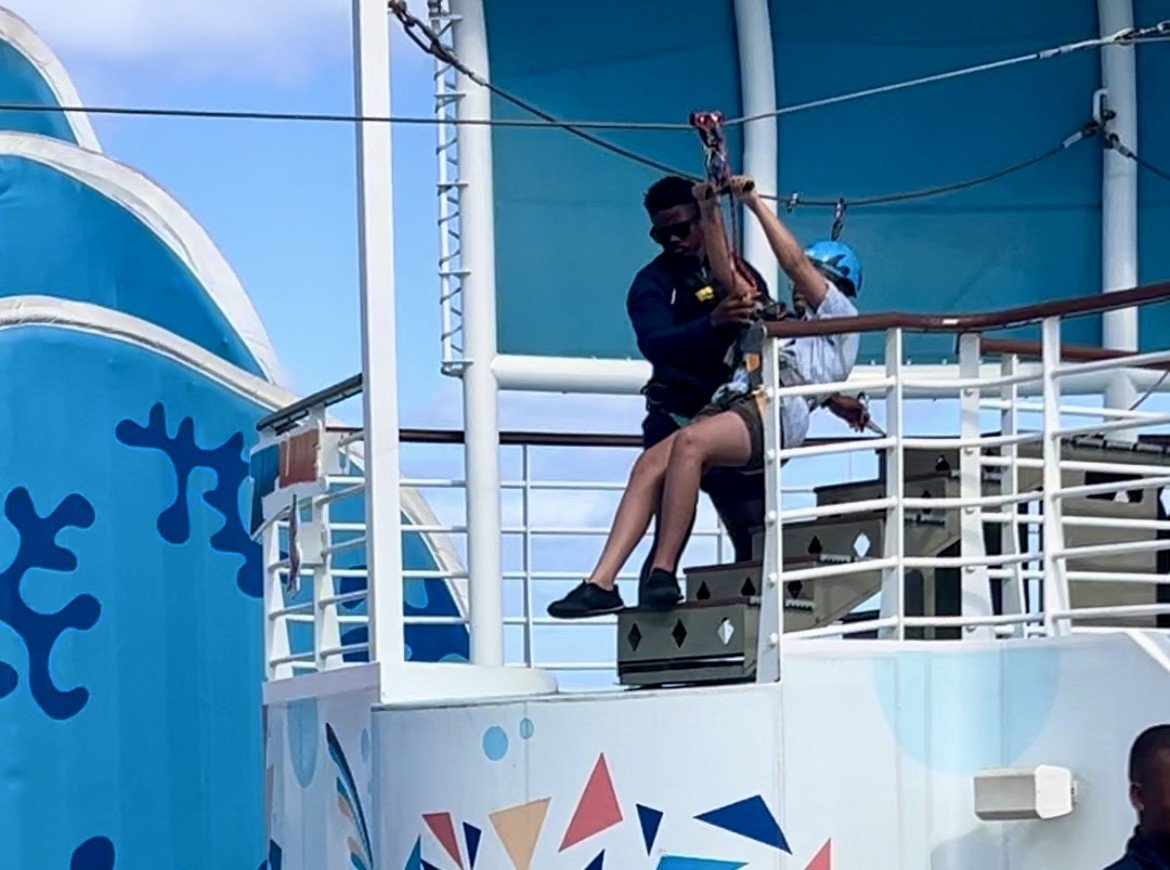 Two people riding a zip line on a cruise ship, with one person in a blue helmet and the other person in dark sunglasses, surrounded by the ship's white and blue structure.