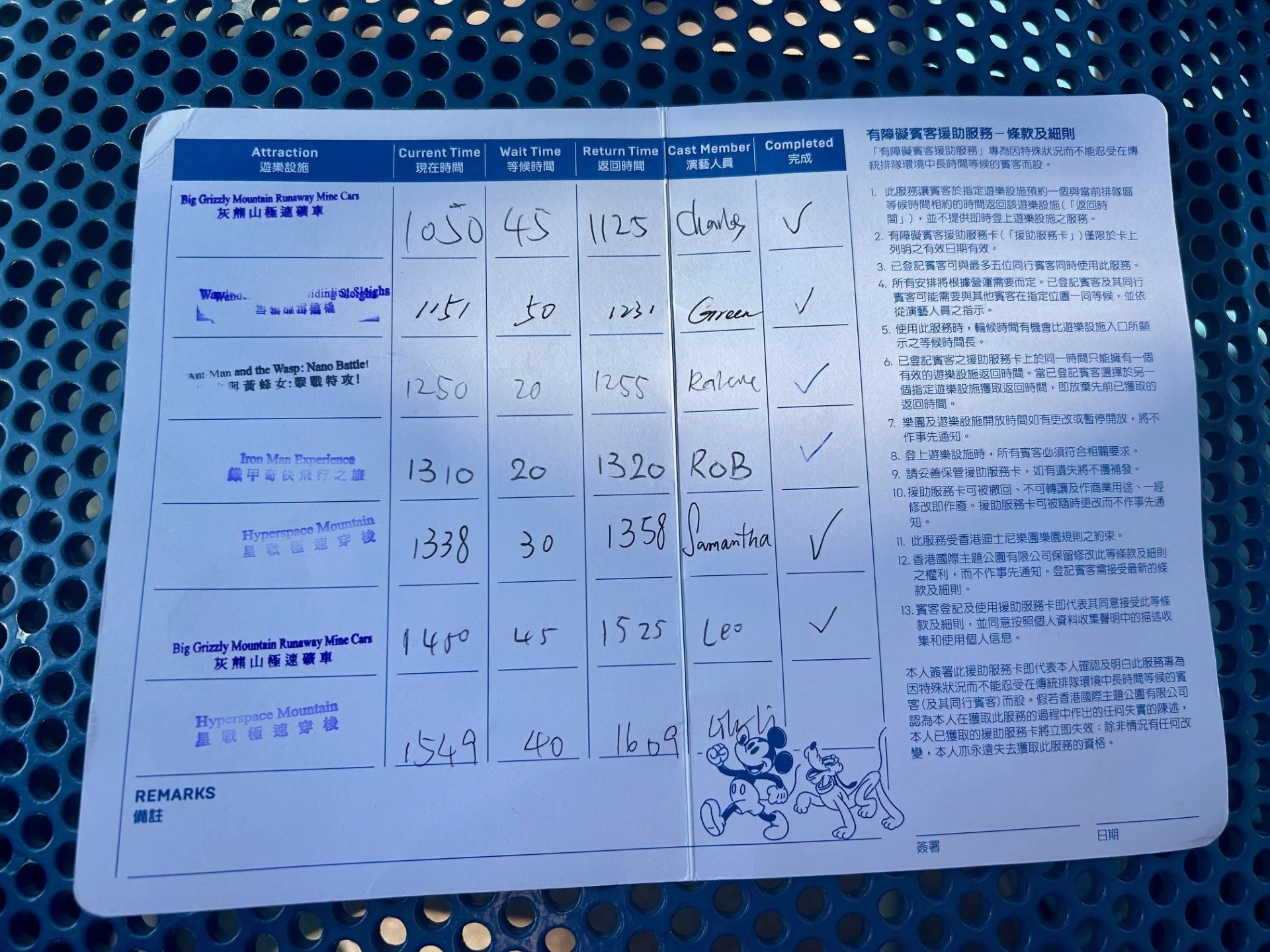 A laminated sheet of paper with a table listing ride information for various amusement park rides, including ride names, current times, wait times, return times, cast members, and completion status. The sheet contains text in both English and Chinese, with some handwritten entries, and features an illustration of Mickey Mouse and Friends at the bottom right corner.