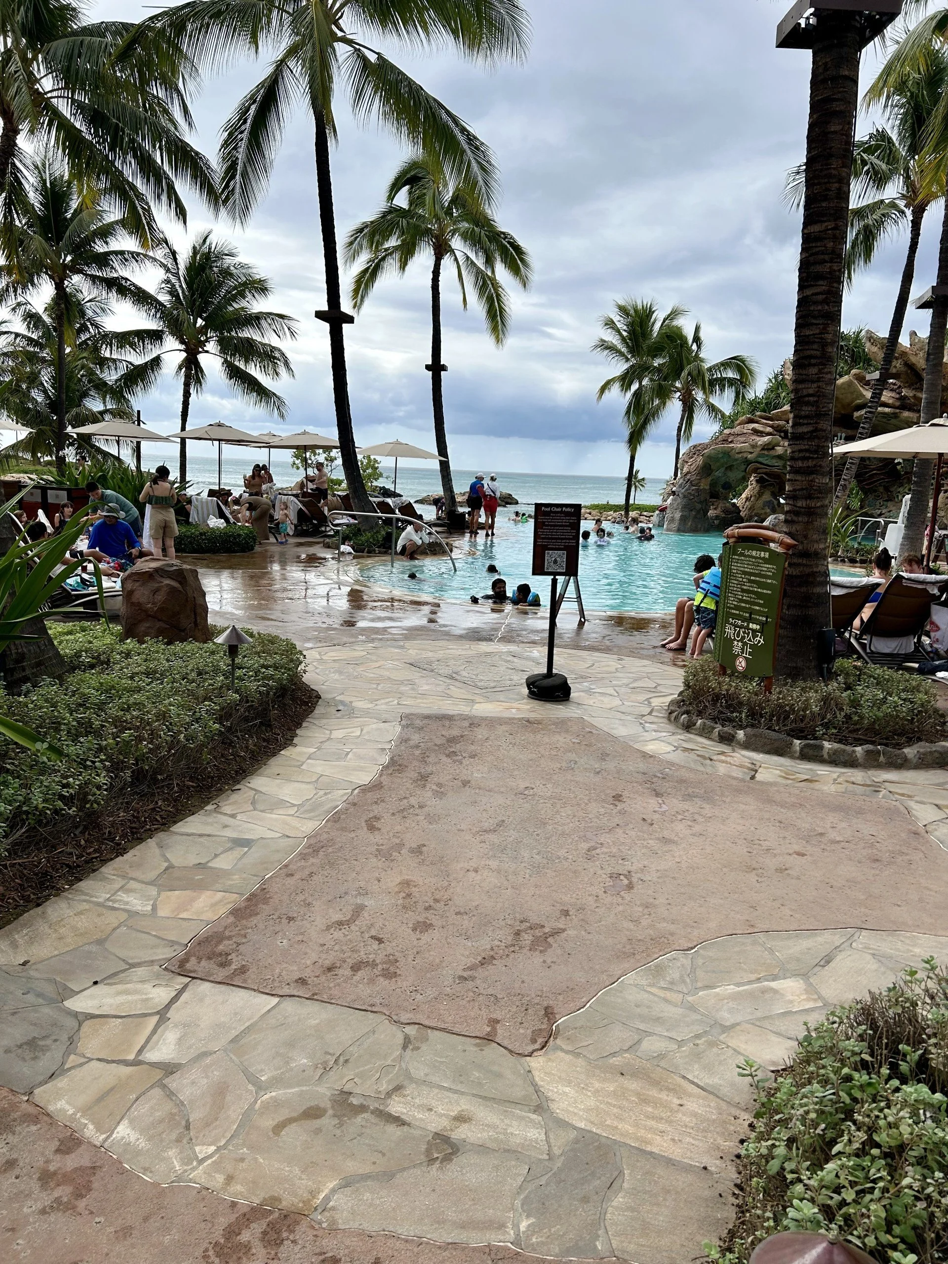 Pool area at a tropical beach resort with palm trees, outdoor lounge chairs, umbrellas, and people swimming and relaxing in the pool. Overcast skies in the background.