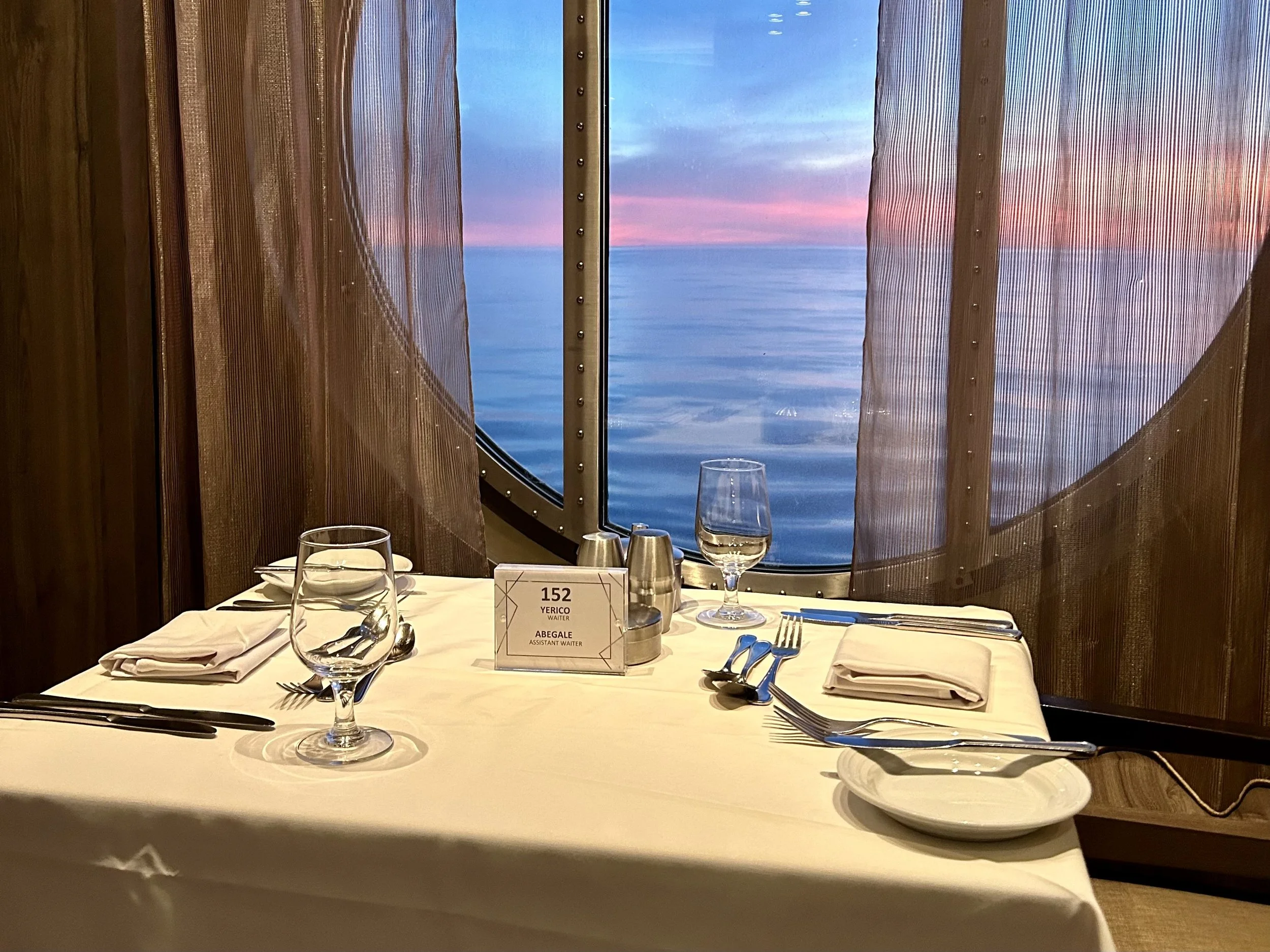 A dinner table set for two in a cruise ship dining room with a large round window showing an ocean and sunset sky, decorated with curtains, glasses, utensils, and a table number sign. Wonder of the Sea Main Dining Room