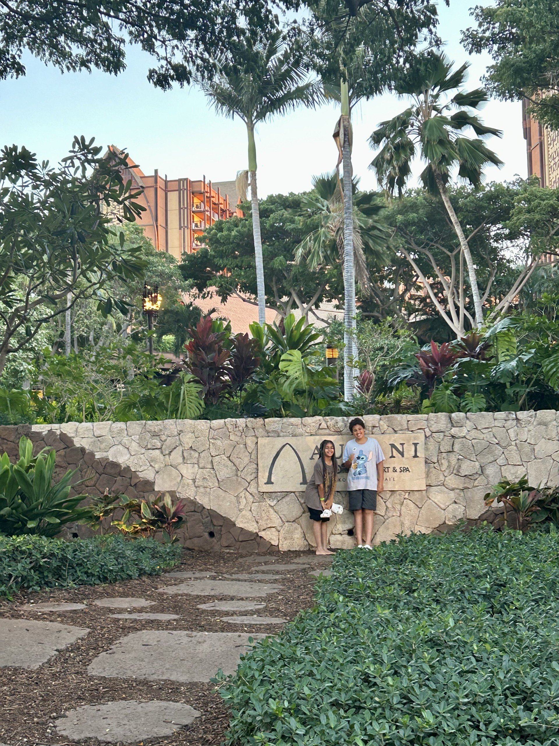 Two people standing in front of a stone sign that reads Aulani, with lush green tropical plants and tall palm trees behind them. The setting appears to be a botanical garden or resort area at Disney's Aulani.