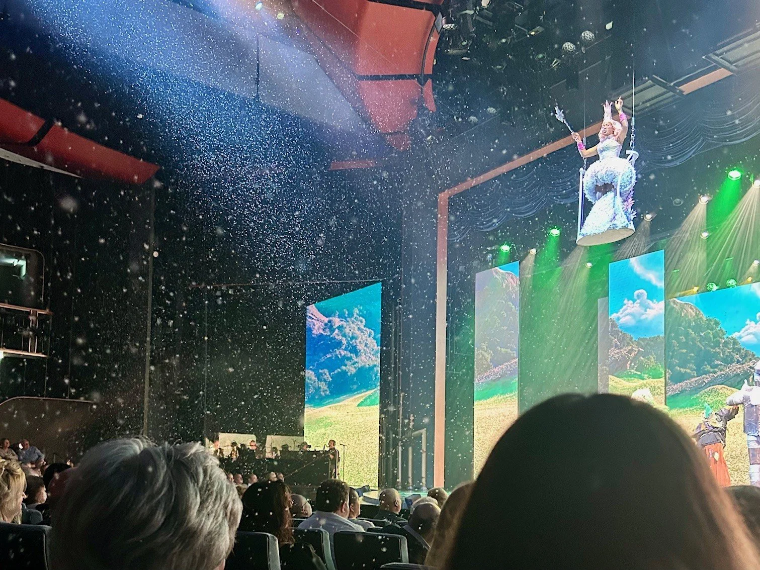 A performer dressed as a fairy on a suspended platform during a circus or stage show, with an audience watching and confetti falling from above.