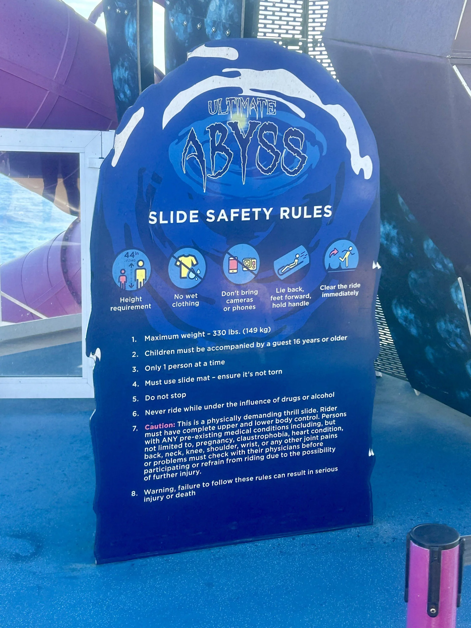 Sign with slide safety rules at an amusement park. It includes rules such as height requirement, no wet clothing, don’t bring cameras or phones, lie back with feet forward, clear the ride immediately, maximum weight 330 pounds, children must be accompanied by an adult, only one person at a time, use a slide mat, do not stop, never ride under the influence, and follow all medical and health guidelines.