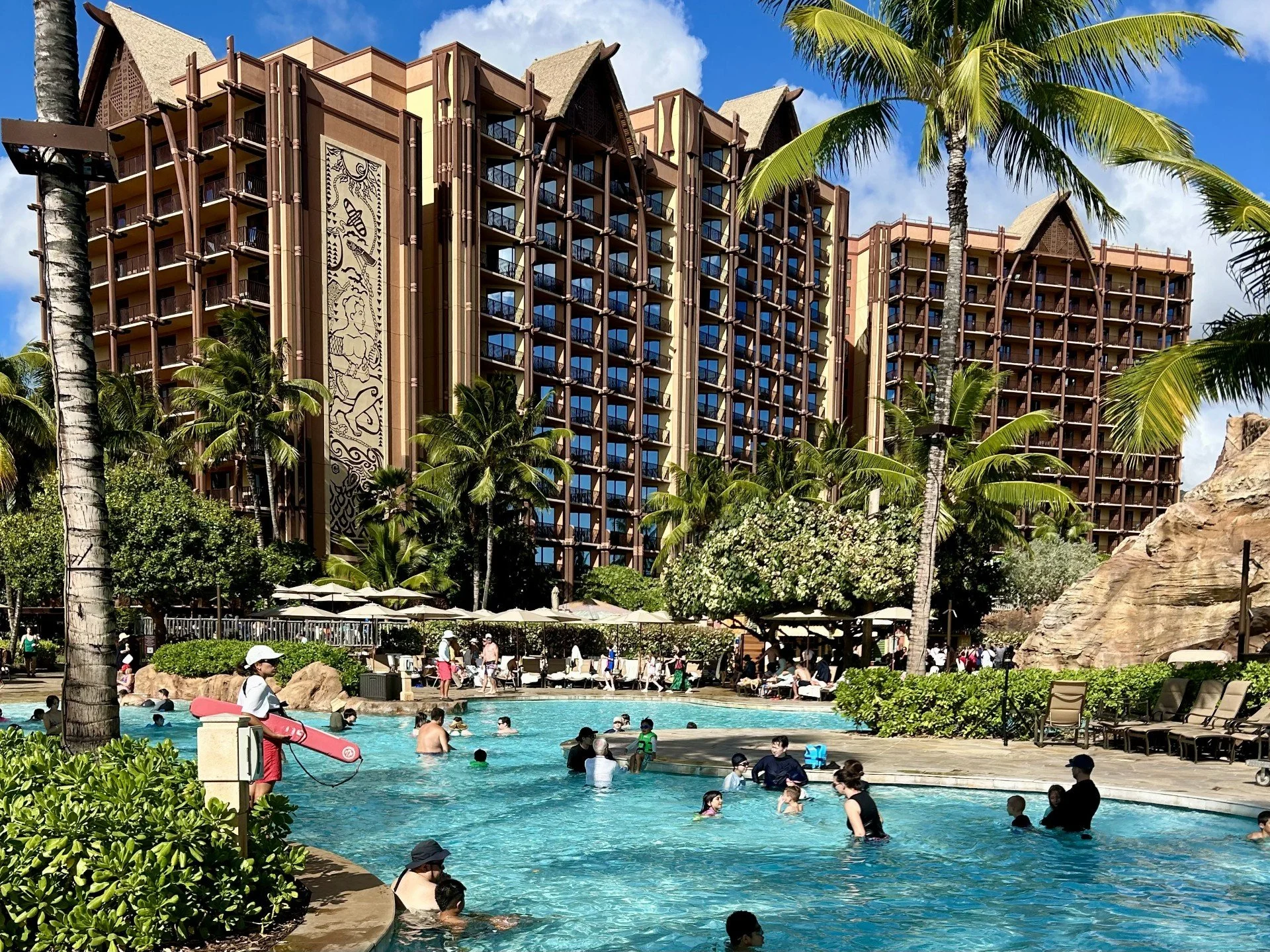 People swimming and relaxing by a resort pool at Disney's Aulani in Oahu Hawaii with tall palm trees and a large multi-story hotel building in the background.