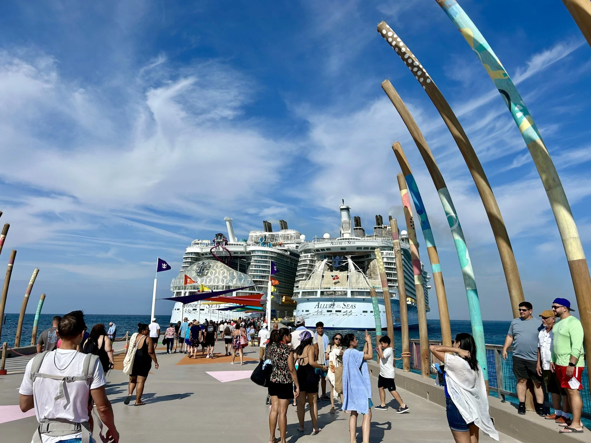People walking along a pier with a large cruise ship in the background on a sunny day.