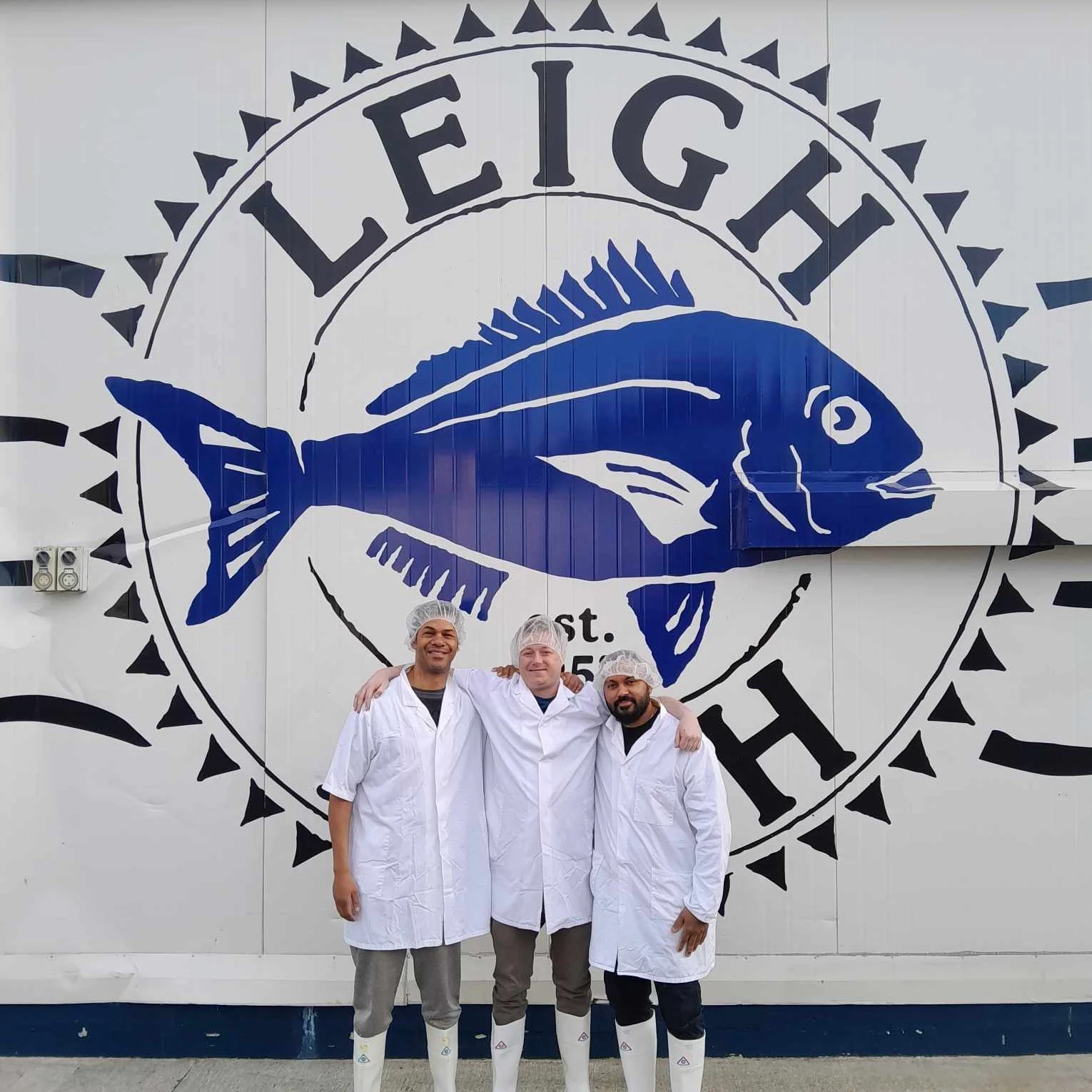 Three men standing under a large sign for Leigh Fisheries.