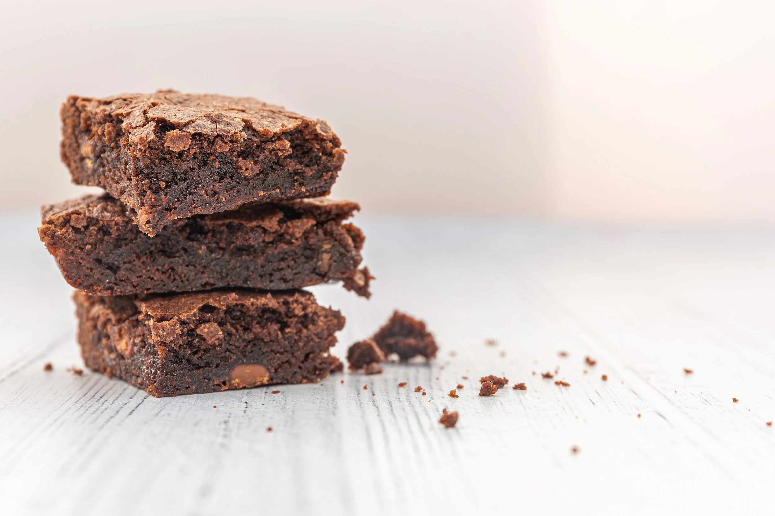 Stack of three chocolate brownies on a white wooden surface with crumbs.