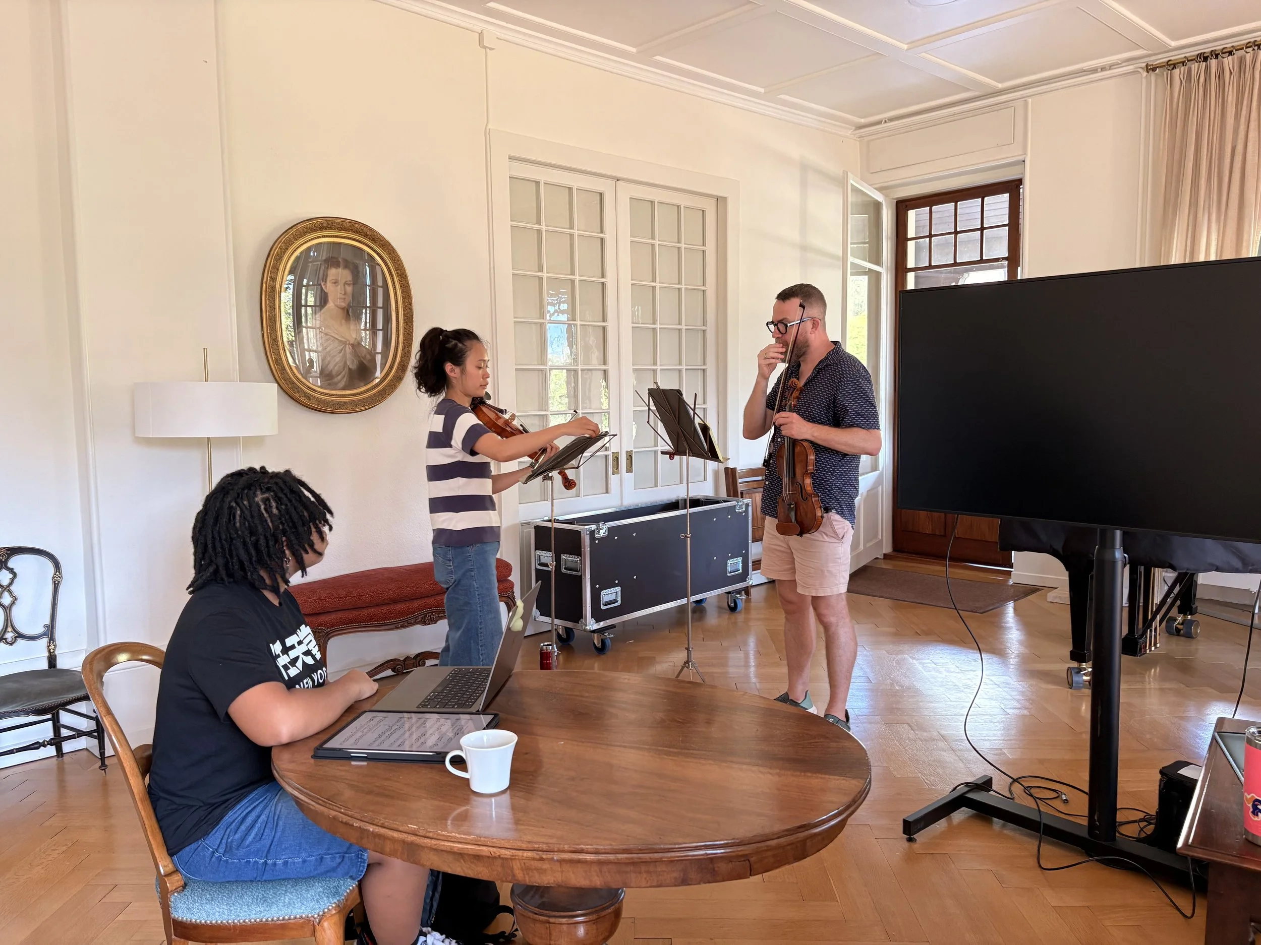 A woman at a table looks on as two violinists, a student and a teacher, discuss their music