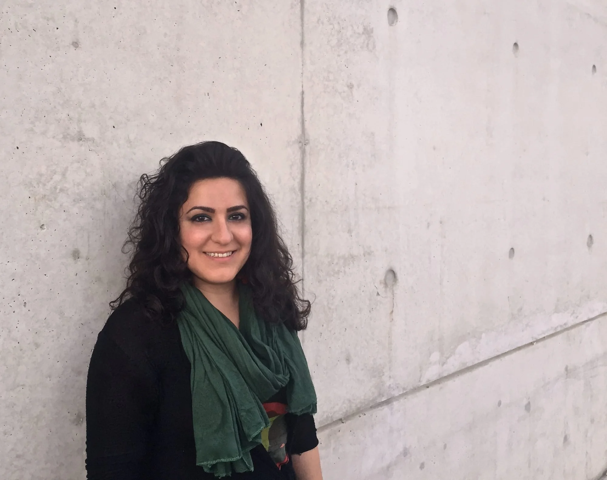 Anahita Abbasi smiles in a green scarf standing in front of a concrete wall