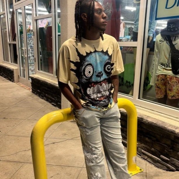 Young man with dreadlocks wearing a graphic t-shirt and light-colored pants, standing outside a store at night with his hands in his pockets, leaning against a yellow barricade.