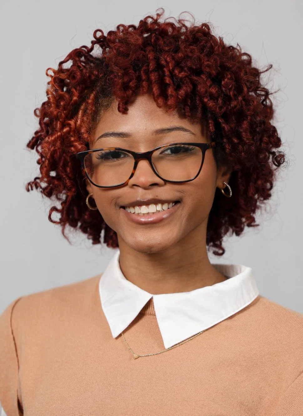 A woman with red curly hair wearing glasses, gold hoop earrings, a peach top, and a white collar shirt, smiling against a plain gray background.
