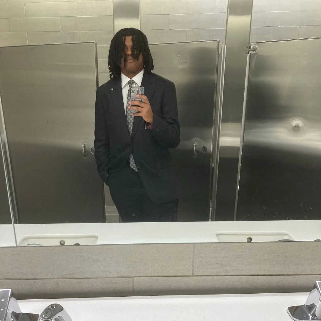 Young man in a black suit taking a mirror selfie in a bathroom with gray tiled walls and stainless steel stalls.