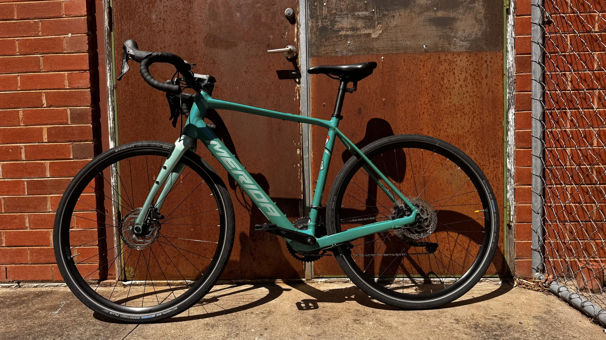 Teal Merida e gravel bicycle with black accents, parked against a brown rusty metal door with a brick wall and chain-link fence on the sides.