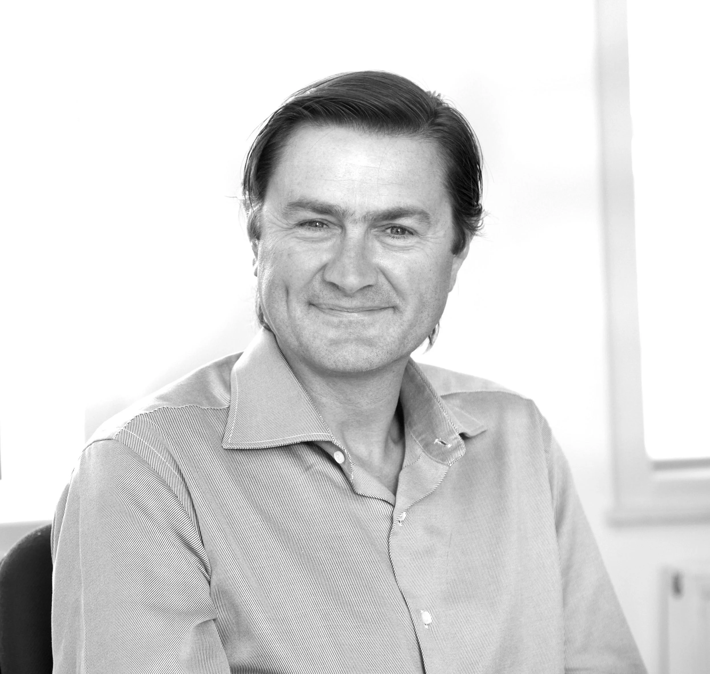 Black and white photo of a smiling man with dark hair, wearing a collared shirt, sitting indoors near a window.