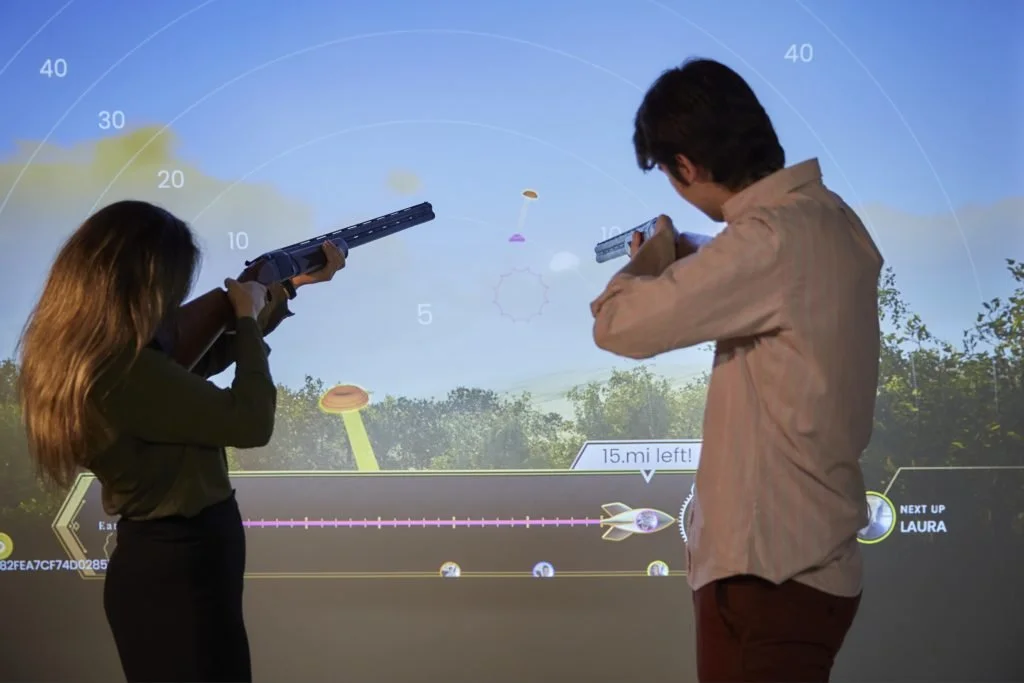 Two people are playing a virtual shooting game using toy guns in front of a large screen displaying a space scene.