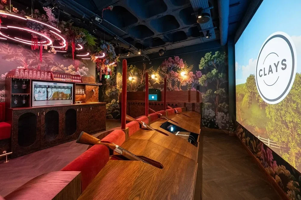 A golf simulation room with a large screen showing the CLAYS logo, holding virtual clay shooting games, surrounded by wooden desks and red upholstered chairs, ambient lighting, and floral wallpaper decor.